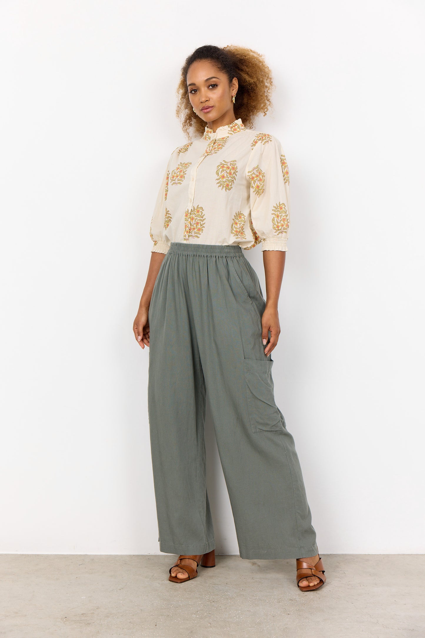 A person poses confidently in SoyaConcept's Linen Blend Pant Ina 64-C 41024-40, paired with a floral-patterned blouse against a white background. With curly hair and sandals, their hand on the hip captures the chic essence of a summer wardrobe.