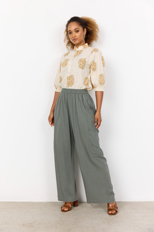 A person poses confidently in SoyaConcept's Linen Blend Pant Ina 64-C 41024-40, paired with a floral-patterned blouse against a white background. With curly hair and sandals, their hand on the hip captures the chic essence of a summer wardrobe.