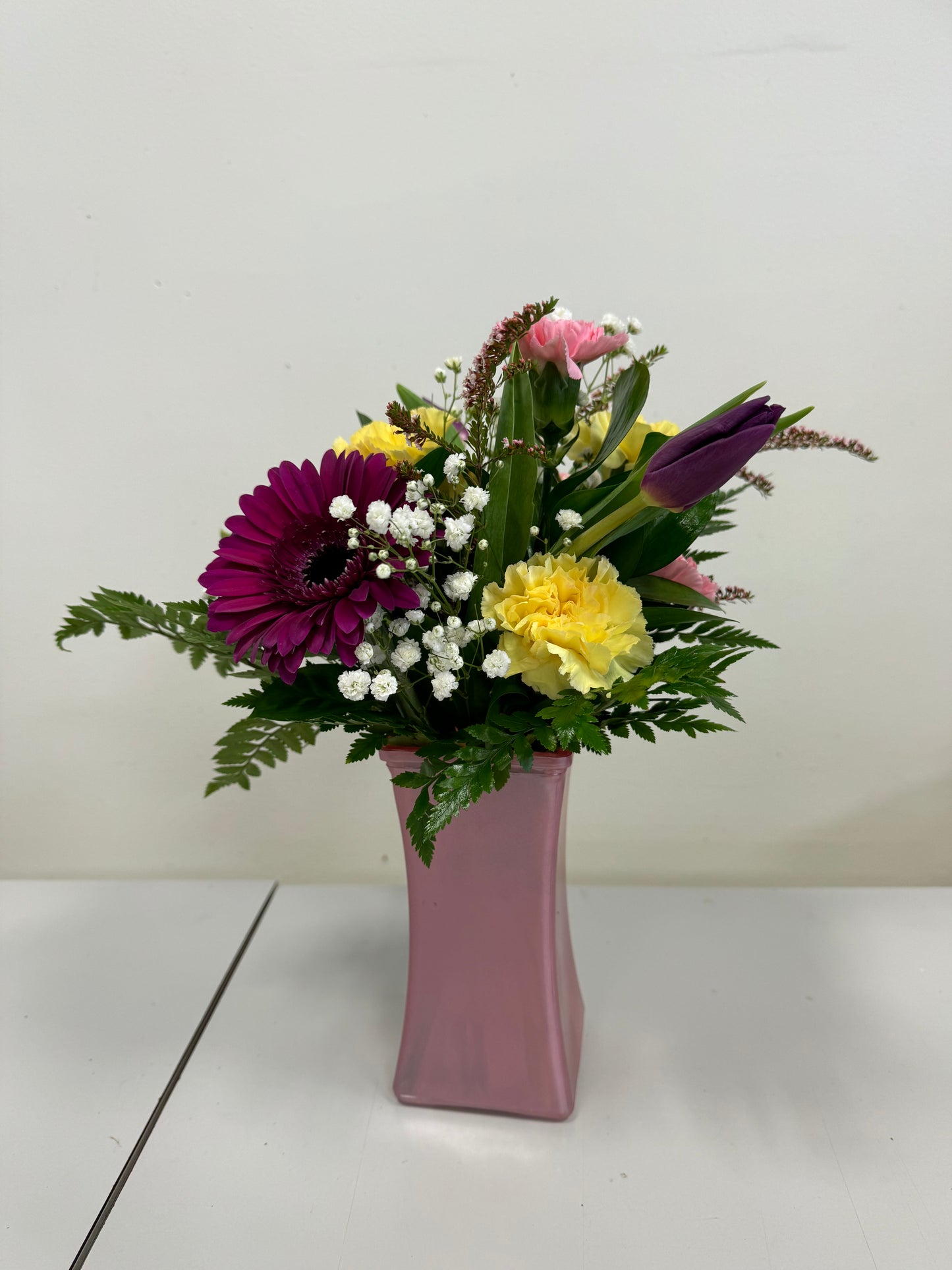 A Designer’s Choice Vase from Posies Flowers & Fashion, filled with white lilies and green foliage, graces a marble table in an elegantly modern room. Soft lighting and contemporary furniture in the background create a warm and inviting atmosphere, showcasing the artistry of floral designers, subject to availability.