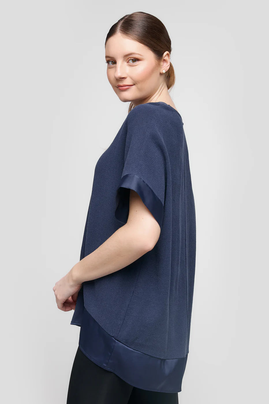 A person with long hair tied back stands sideways, smiling over their shoulder. They're wearing the loose-fitting TOP CS25-609 UCHUU Montreal dark blue blouse with satin trim and black pants. The background is a plain light gray.