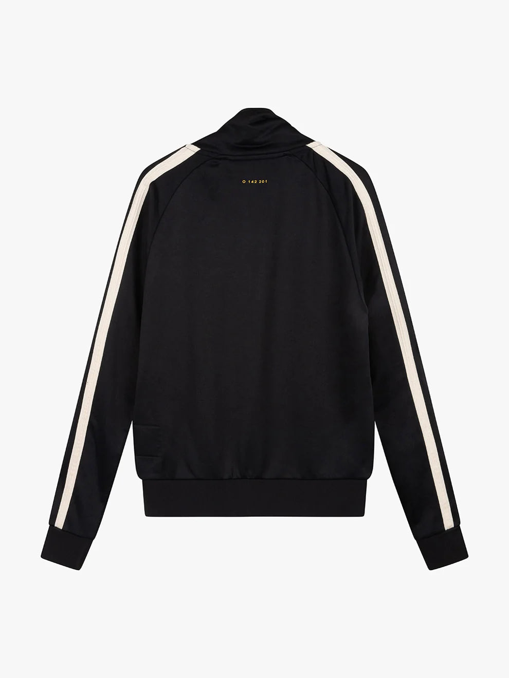 A person with short, curly hair stands in a dark, minimalist studio wearing the Once We Were Warriors Haku Track Jacket - Black and matching pants, which are both adorned with white stripes down the sleeves and legs. The sportswear set features a circular emblem on the chest and thigh. They are also wearing black sneakers and looking downward.