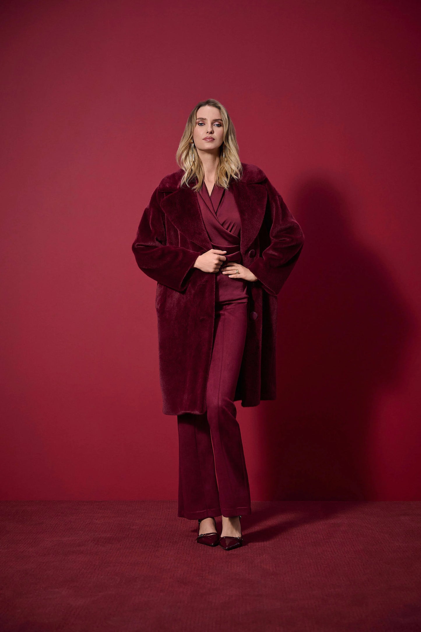 A woman stands against a deep red background, wearing the Joseph Ribkoff Faux Fur Straight Coat 243902F25 with a notched collar over matching flared pants and pointed heels, looking confidently ahead.
