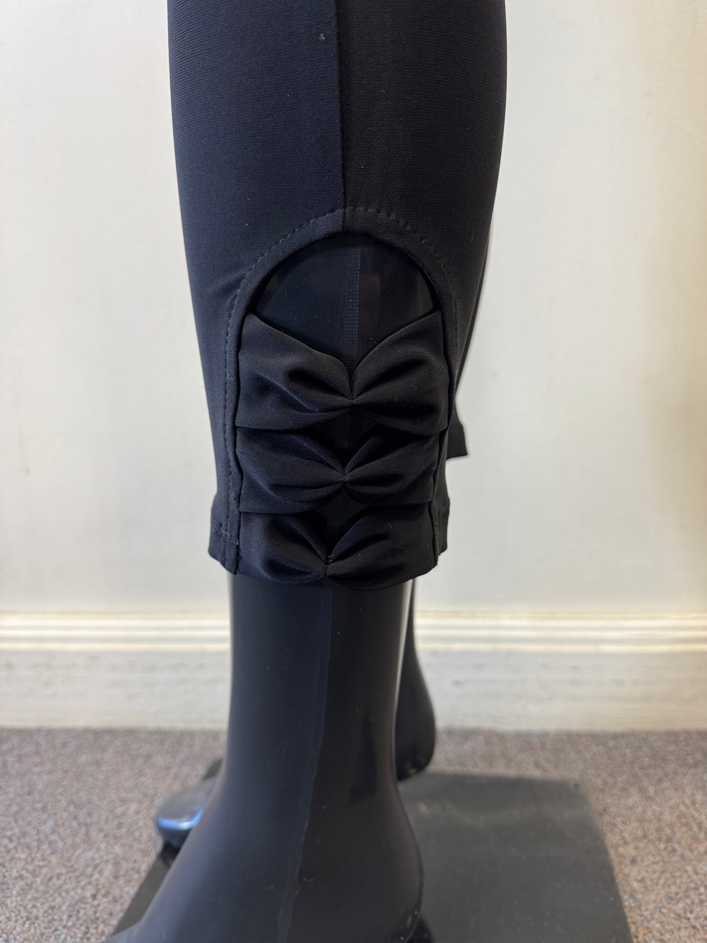 A close-up of a sleek black mannequin leg wearing Bali Corp's Pant 8622, crafted from polyester and spandex. These elegant leggings feature decorative bows on the lower cuff, enhancing the sleek design. The mannequin stands on a square base against a pale wall.