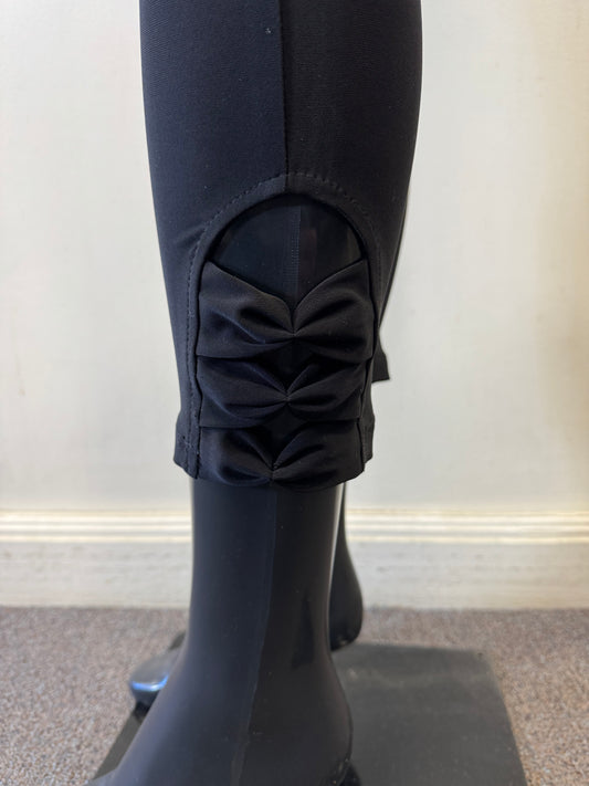 A close-up of a sleek black mannequin leg wearing Bali Corp's Pant 8622, crafted from polyester and spandex. These elegant leggings feature decorative bows on the lower cuff, enhancing the sleek design. The mannequin stands on a square base against a pale wall.
