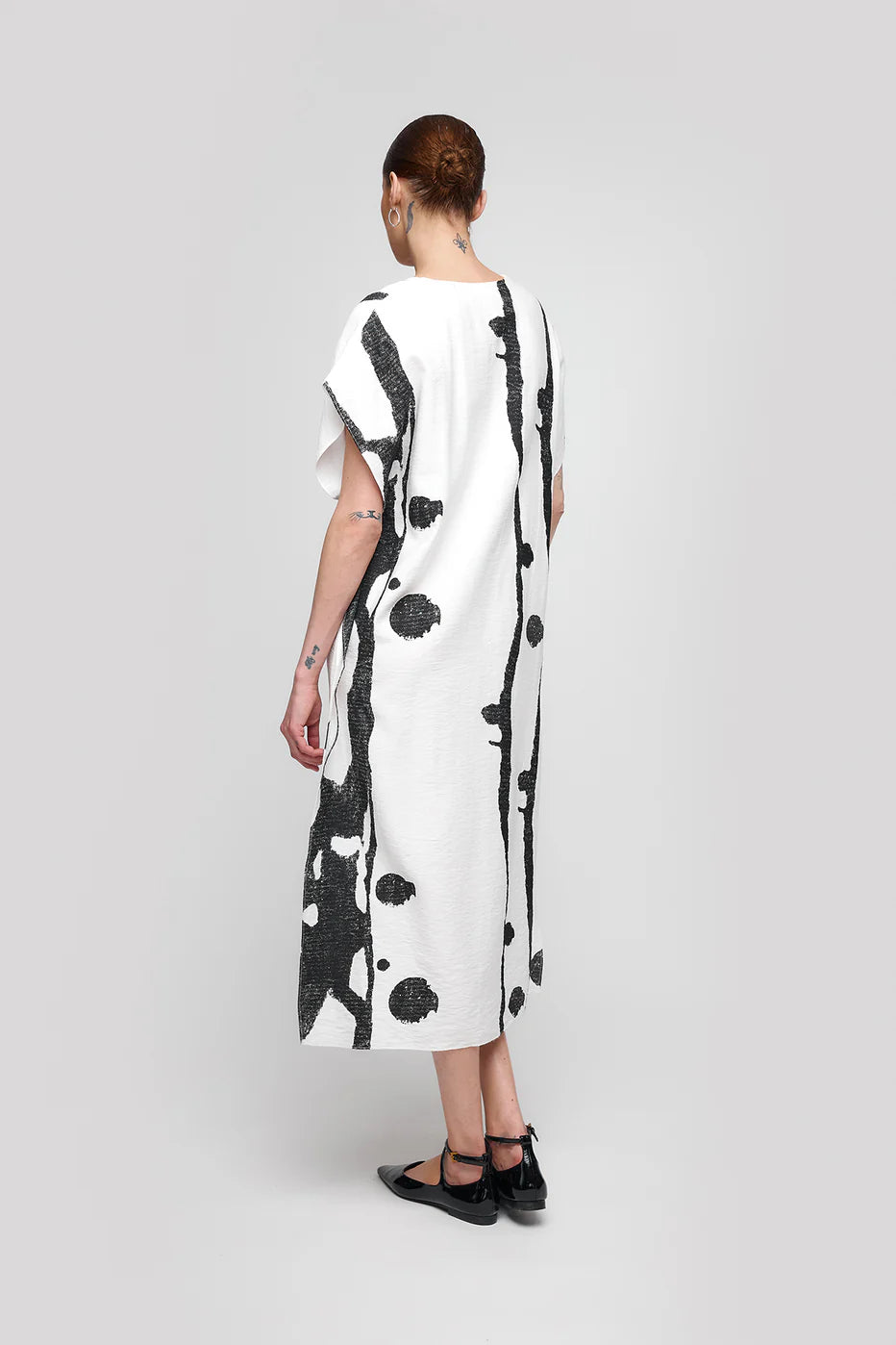 A person with short, tied-back hair wears the UCHUU PRINTED SLEEVELESS SQUARE DRESS CS25-026, showcasing abstract black patterns and a loose fit. They stand on a neutral background facing away to complete the look with black flat shoes.