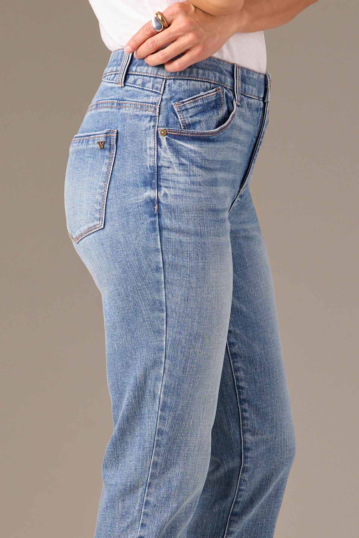 A person models stylish Logan Hi-Rise Skinny jeans in light blue by Wit & Wisdom, paired with a white top. From the side view, they rest their left hand on their hip, displaying a ring on their finger and showcasing the comfortable fit of these P2811YB1 LBA jeans.