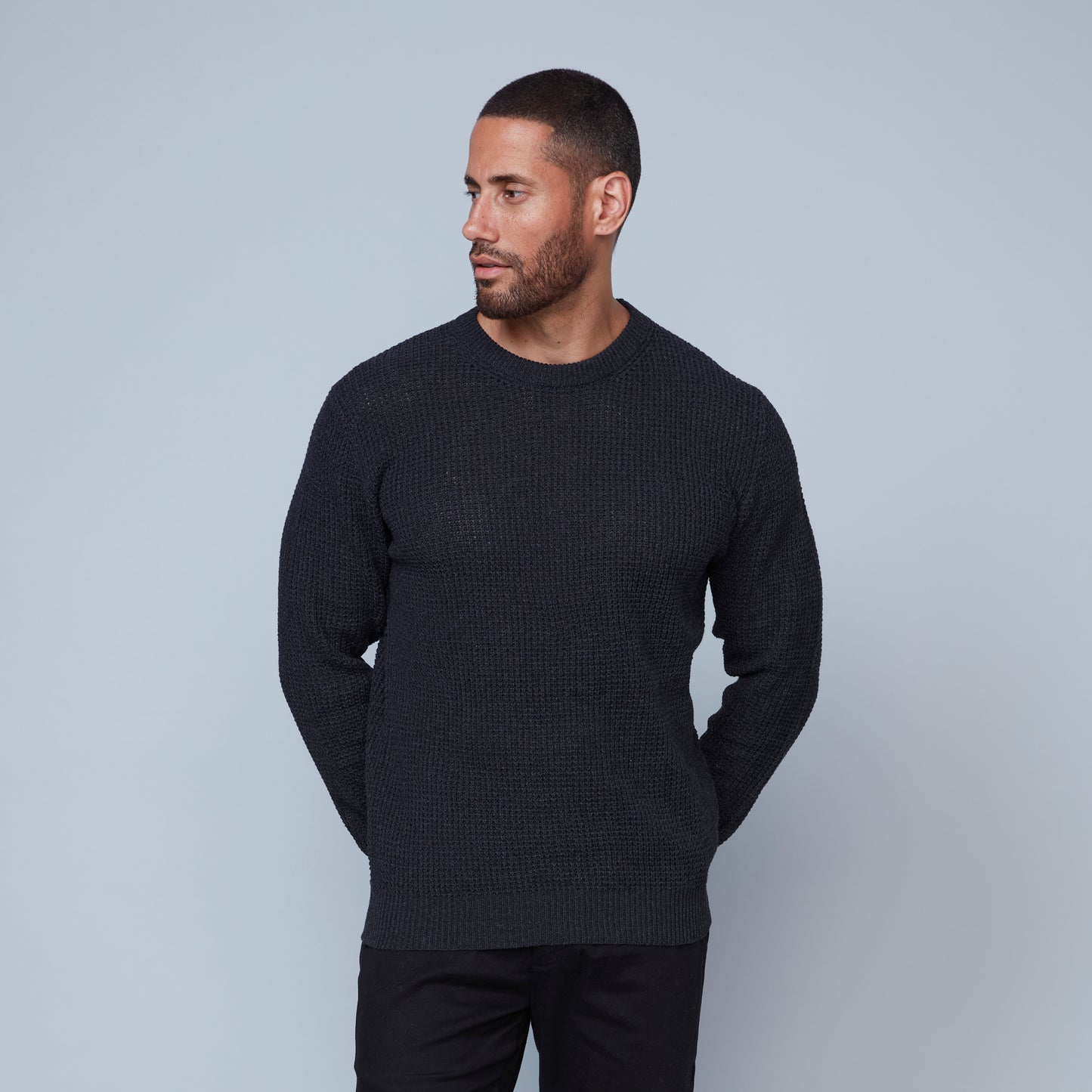 A man with short hair and a beard stands against a plain gray background. He is wearing a dark gray, long-sleeved Crewneck Sweater from HEDGE, made from recycled cotton and black pants, looking slightly to his left. His right arm hangs by his side, and his left arm is bent at the elbow.