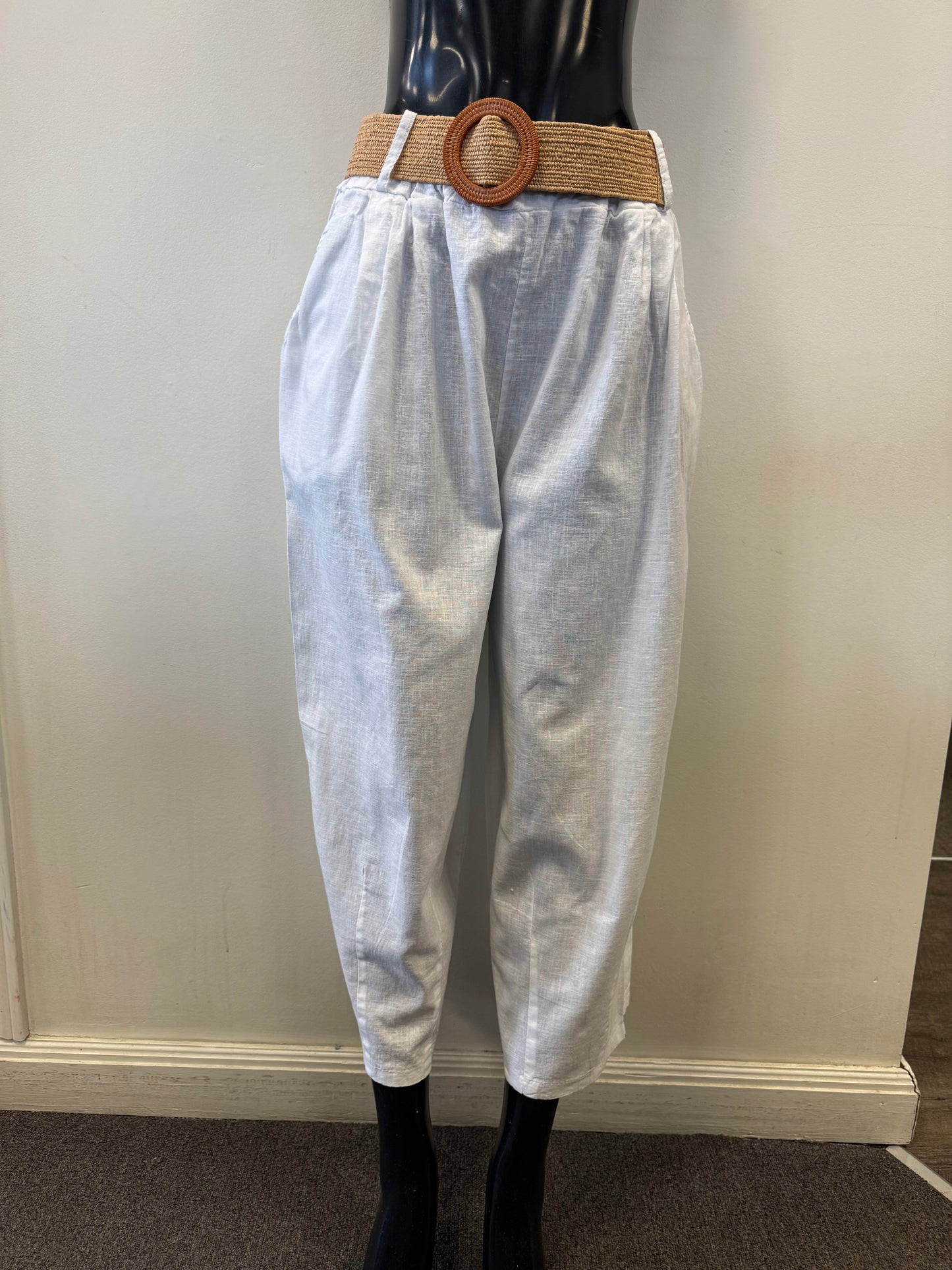 A mannequin displays loose-fitting, light grey Pants with Belt LX38430 by Paolo Tricot, made from a cotton-linen blend for comfort. These high-waisted pants include a tan braided belt with a large circular buckle, set against a simple background of plain walls and carpeted flooring.
