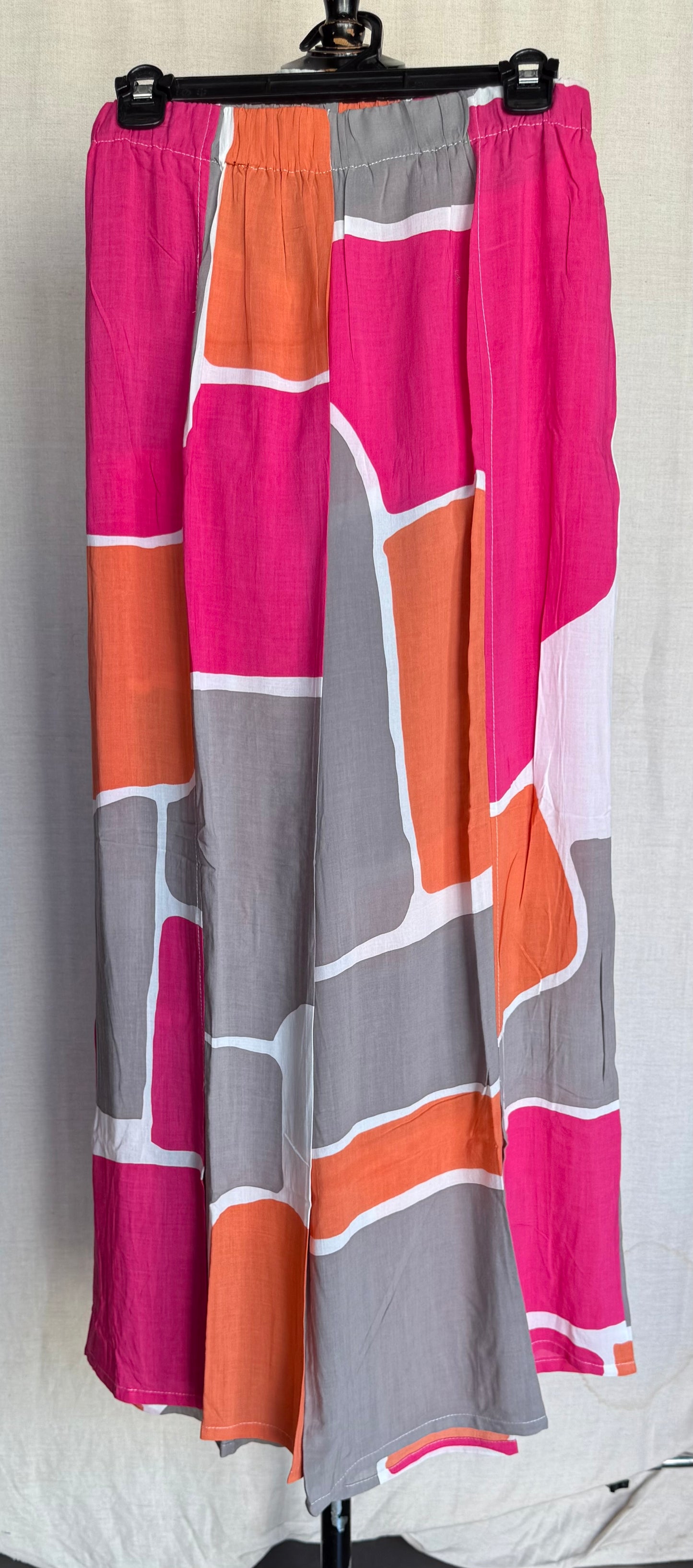 The TaraVao Pant Maora 4016 features a bold geometric pattern in pink, orange, gray, and white on lightweight rayon, displayed on a black hanger against a light background.