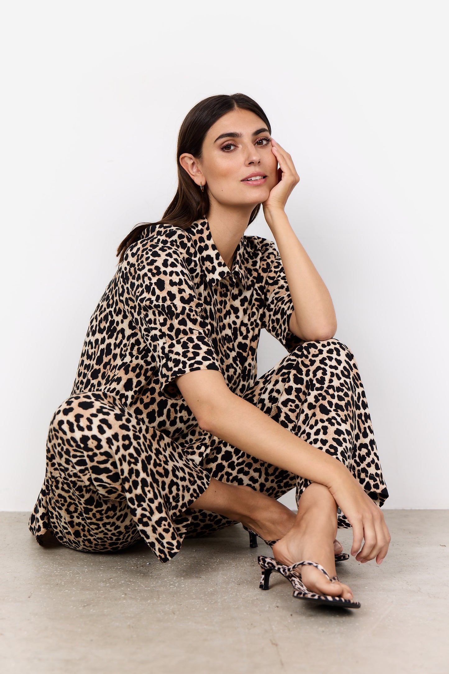 A person poses on a white background wearing the luxurious animal print Button Shirt Pietta 2 by SoyaConcept, matching heels, and sits on the floor with one knee up, resting their chin on their hand while directly gazing at the camera, exuding a fierce Pietta 2 vibe.