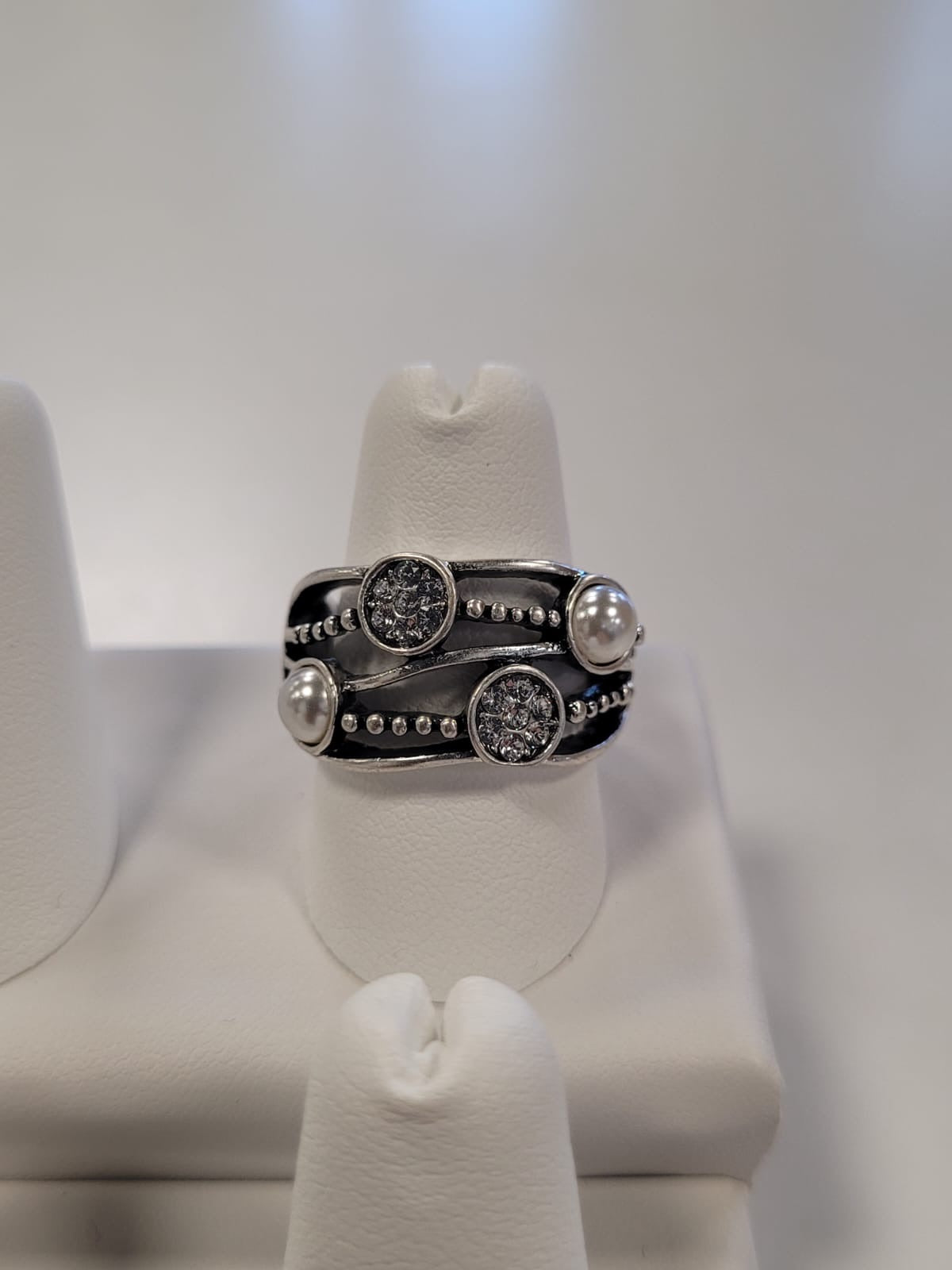 A stunning Jenna-61 Adjustable Ring by Elise Accessories, showcasing a design with three circular, CZ stone-studded elements and two white pearls, displayed on a white ring stand against a soft, blurred background. The silver ring exudes a modern and elegant appearance with intricate detailing.