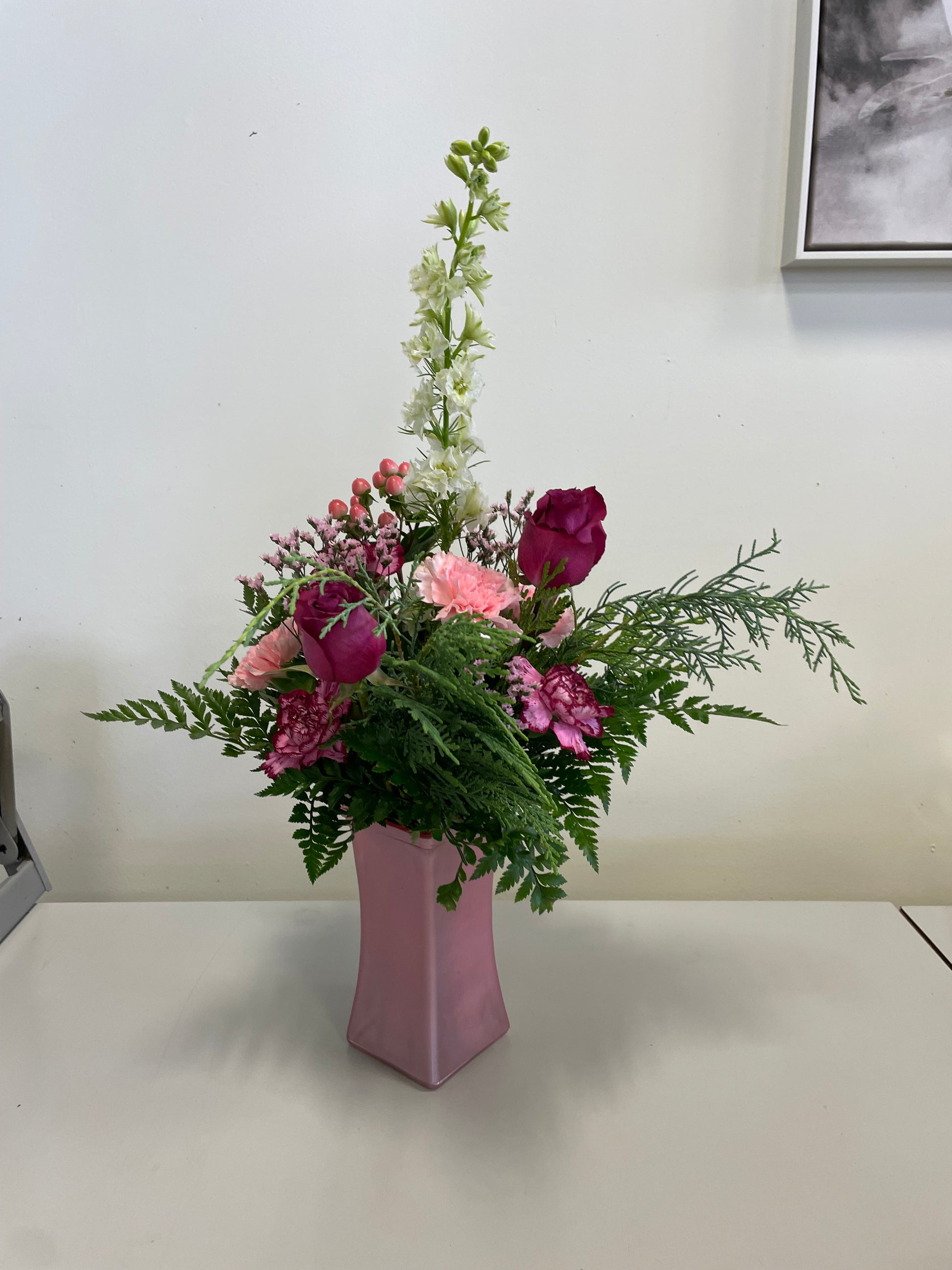 A Designer’s Choice Vase from Posies Flowers & Fashion, filled with white lilies and green foliage, graces a marble table in an elegantly modern room. Soft lighting and contemporary furniture in the background create a warm and inviting atmosphere, showcasing the artistry of floral designers, subject to availability.