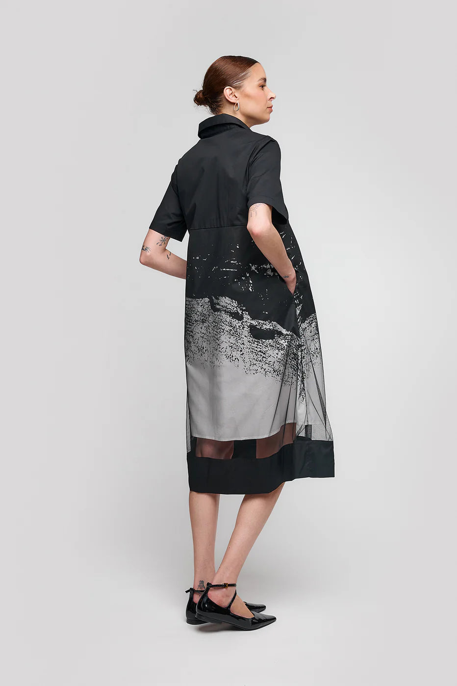 A person with short brown hair showcases the stylish and comfortable SHORT SLEEVE SHIRT DRESS WITH TROMPE L'OEIL PRINT by UCHUU, featuring a graphic design and flowy silhouette. They stand sideways in black flat shoes against a plain gray background.