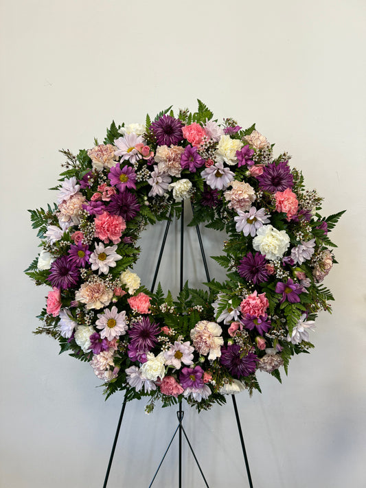 The "Soft Embrace Wreath" by Posies Flowers & Fashion is a large, circular wreath on a stand, beautifully adorned with a vibrant mix of purple, pink, and white flowers in full bloom, and accented with lush green foliage. It is displayed against a plain, light-colored wall. Please note that availability may vary based on seasonality.