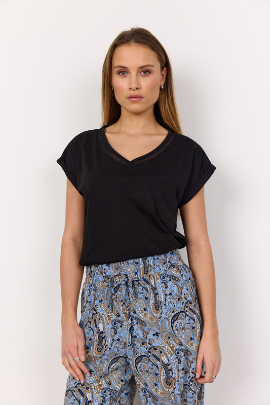 A person with long hair wears a black V-neck shirt ("V-neck Shirt Babette 57 SoyaConcept") made from sustainable fabric and blue paisley pants against a white background, maintaining a neutral expression while looking at the camera.