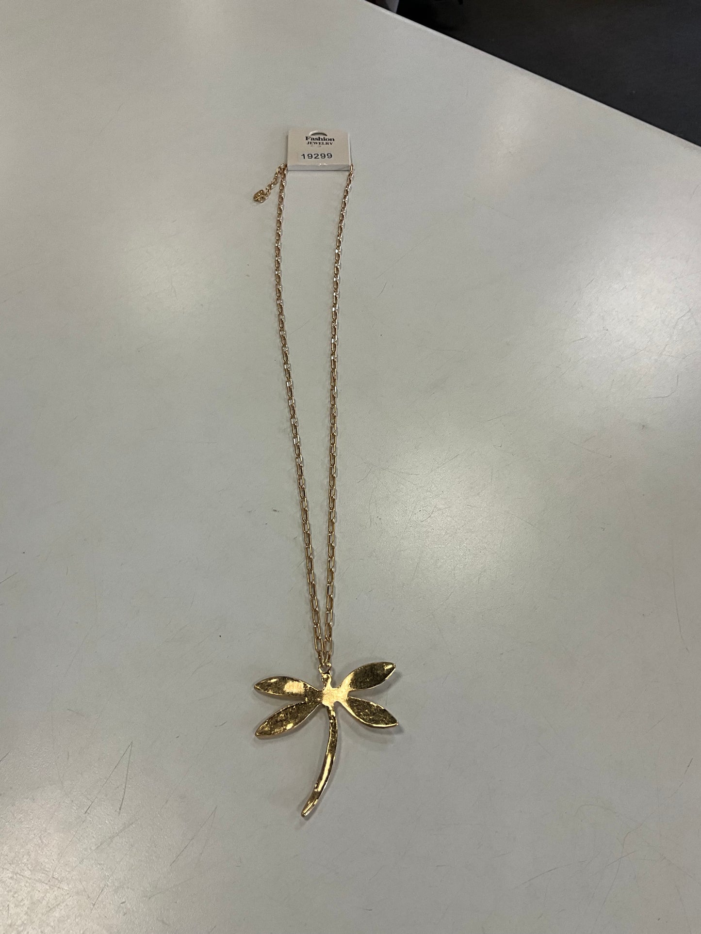 The Ava 22 Gold Necklace from Elise Accessories, featuring a hammered dragonfly-shaped pendant, is displayed on a white surface with a price tag attached to the chain near the top.
