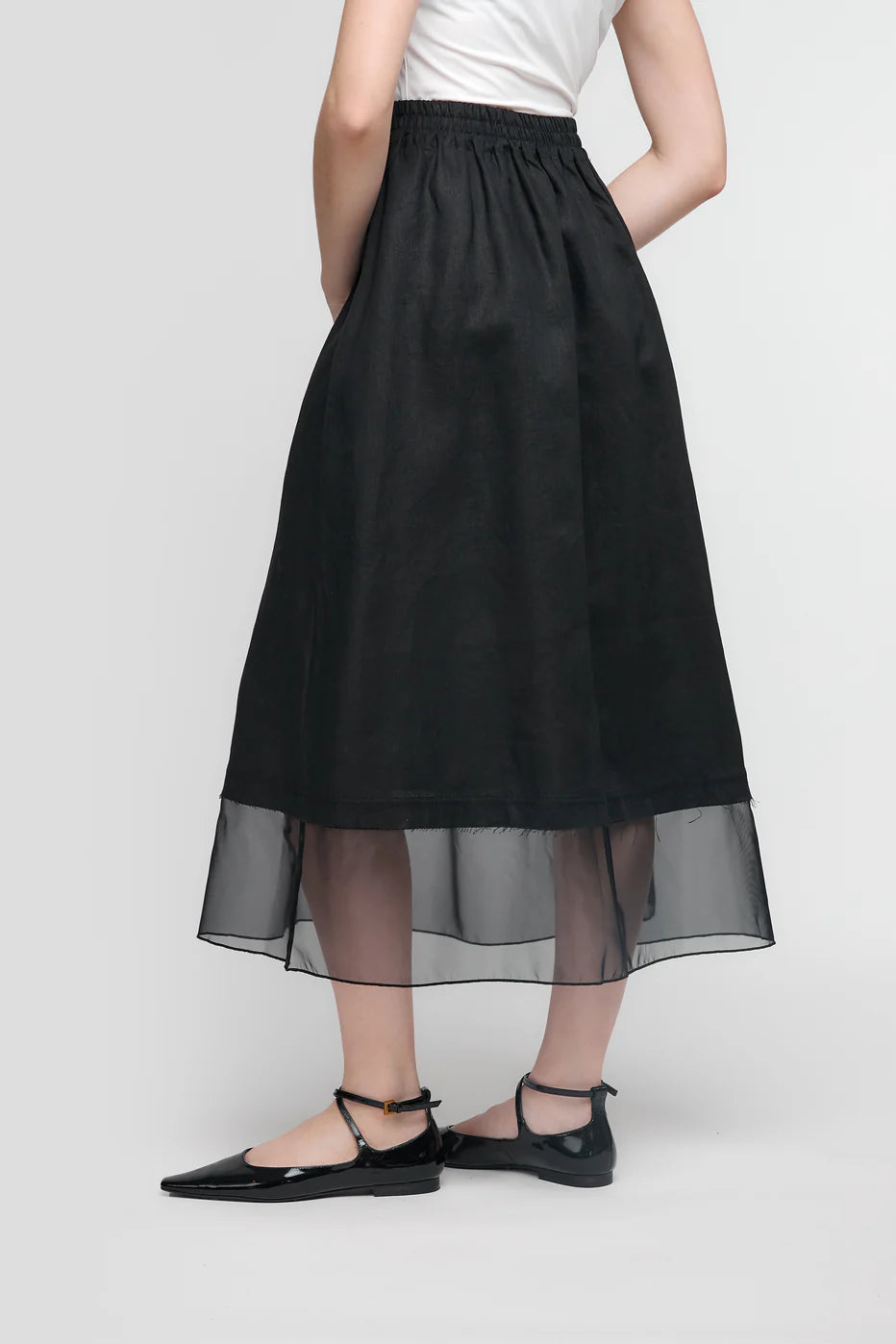 A person in a white top and the UCHUU SKIRT WITH MESH PETTICOAT, CS25-203, stands with their back to the camera. They wear black pointed flats with ankle straps in a simple, light setting that highlights UCHUU Montreal's minimalist elegance.