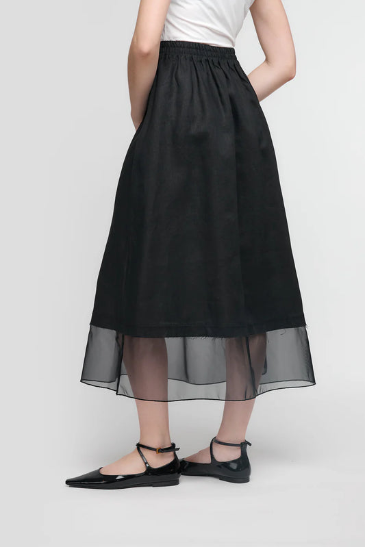 A person in a white top and the UCHUU SKIRT WITH MESH PETTICOAT, CS25-203, stands with their back to the camera. They wear black pointed flats with ankle straps in a simple, light setting that highlights UCHUU Montreal's minimalist elegance.