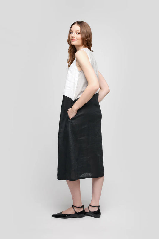 A person with long hair smiles, wearing the TWO-TONE SLEEVELESS TRAPEZE DRESS CS25-020 from UCHUU, featuring side pockets. They stand sideways in black ankle-strap flats against a light grey background.
