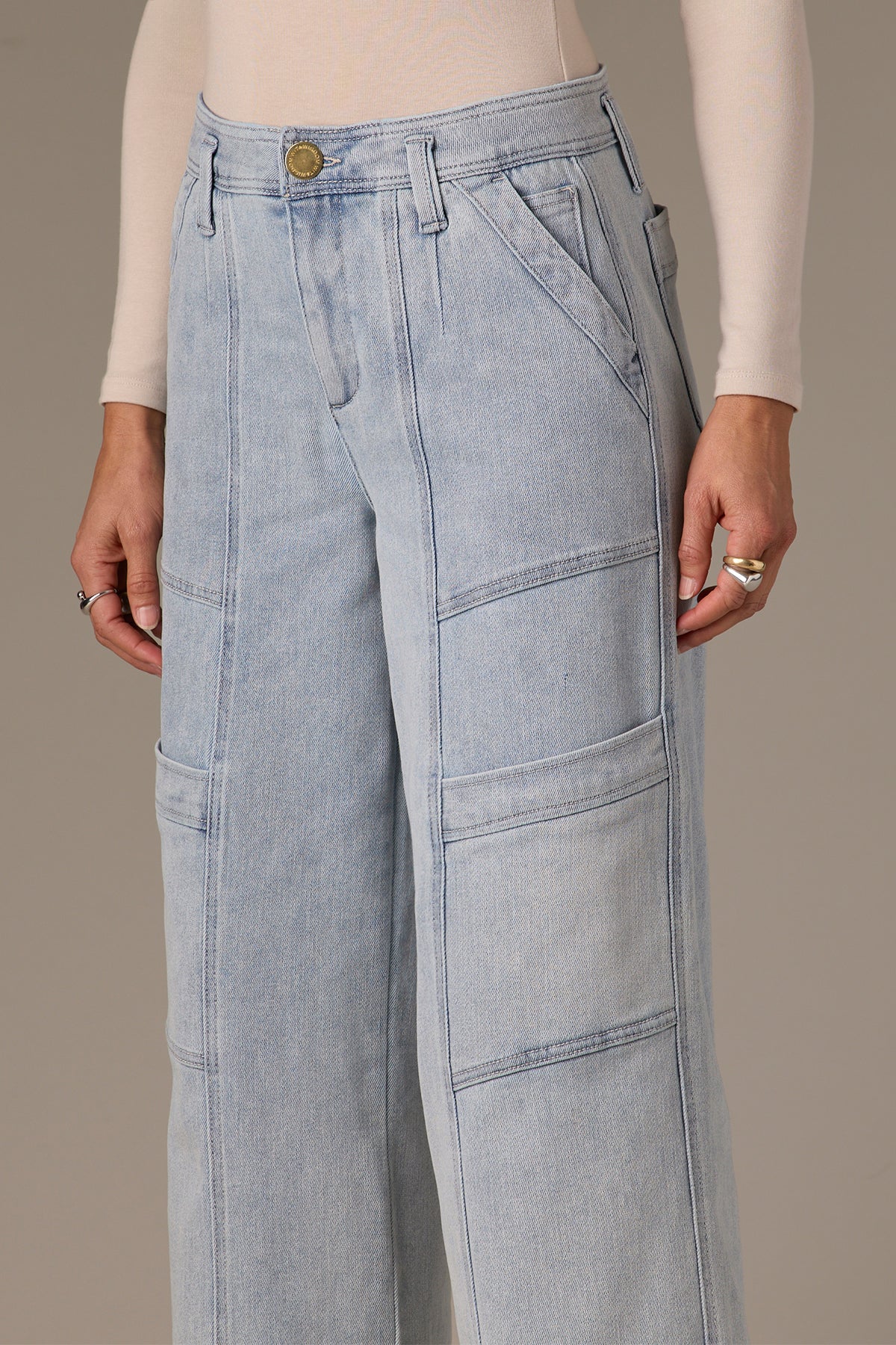 A person wears Wit & Wisdom's Skyrise Double Bottom Wide Leg Jeans in light blue with a long-sleeve beige top. The jeans offer a sustainable, stylish look featuring multiple pockets. Rings on their fingers add flair against the neutral background, enhancing the clothing's details.