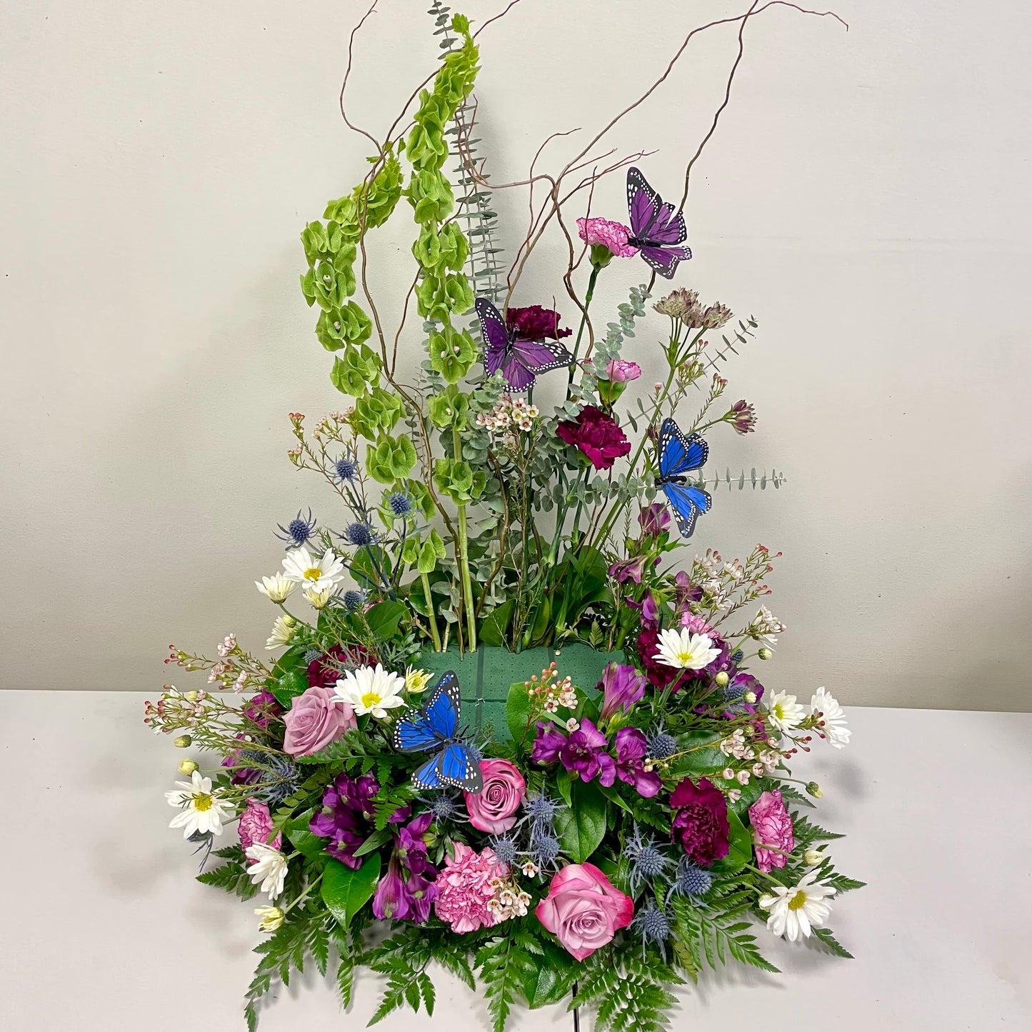 The Garden Memories arrangement from Posies Flowers & Fashion showcases a vibrant floral display featuring pink and purple roses, white daisies, green foliage, and blue butterflies. Enhanced with tall, curly branches and green hanging elements, this elegant and dynamic composition is perfect for adorning a cremation urn.