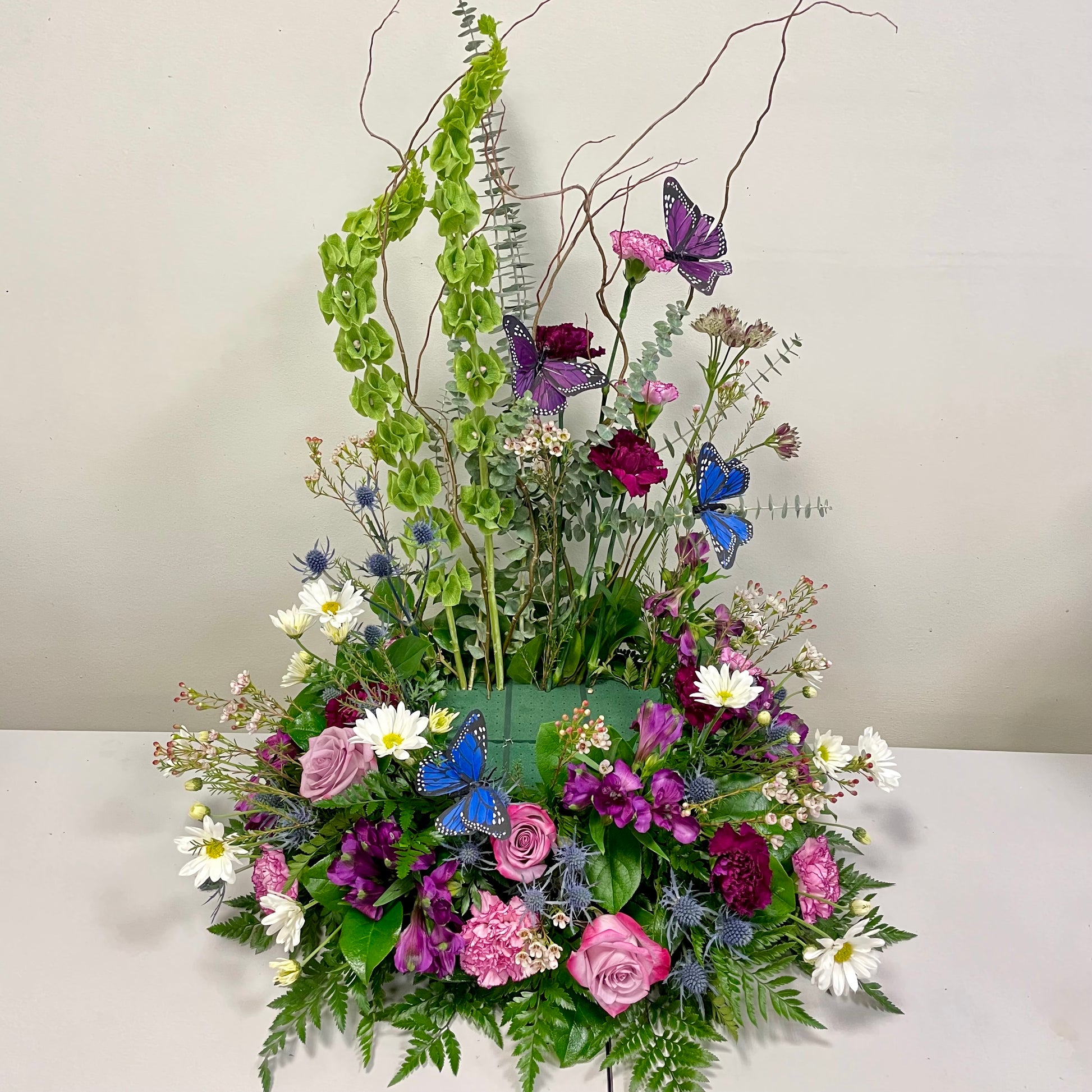 The Garden Memories arrangement from Posies Flowers & Fashion showcases a vibrant floral display featuring pink and purple roses, white daisies, green foliage, and blue butterflies. Enhanced with tall, curly branches and green hanging elements, this elegant and dynamic composition is perfect for adorning a cremation urn.