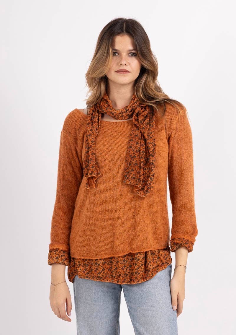 A woman stands against a plain white background, modeling the “Tunic with scarf 1111 VINTAGE: ABRICOT” by LINEN & COTTON HOUSE. She wears the vibrant orange tunic layered over a floral blouse, paired with the matching Abricot scarf. Her long, wavy brown hair cascades over light blue jeans, creating an effortlessly chic look reminiscent of a vintage Italian-inspired ensemble.