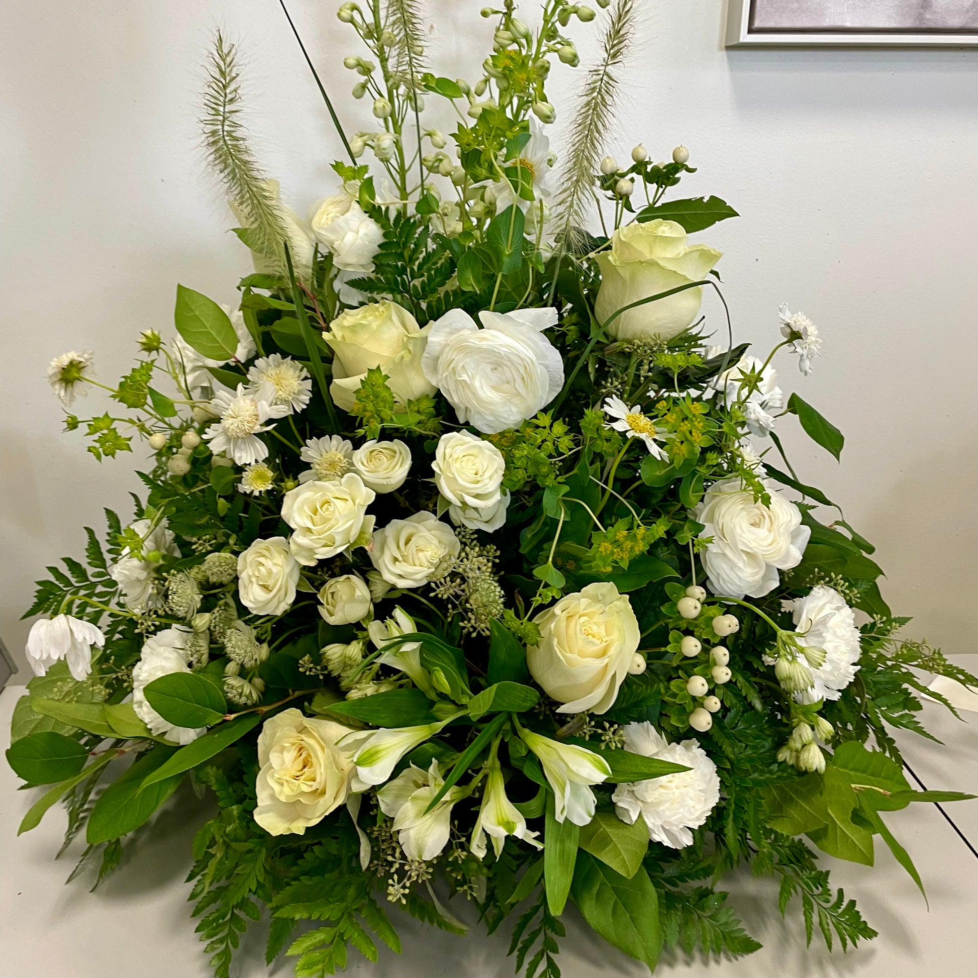 The "Whispering Elegance" by Posies Flowers & Fashion is a grand funeral tribute showcasing white roses, lilies, and assorted green foliage. These delicate flowers are meticulously arranged in an elegant, symmetrical design that results in a lush and full display. The backdrop features a white wall with a partially visible framed picture.