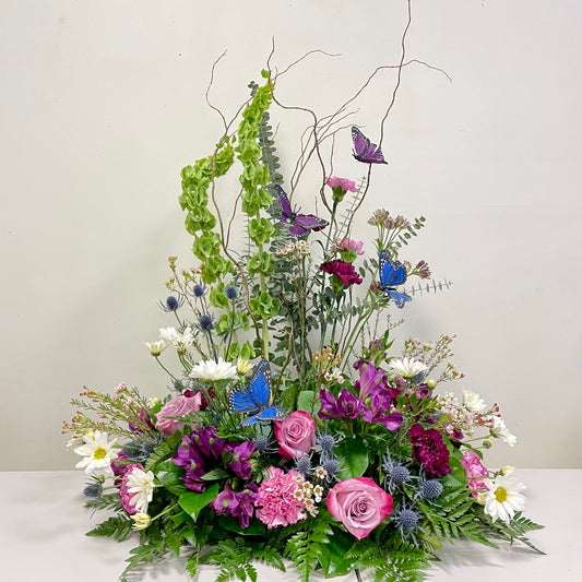 The Garden Memories arrangement from Posies Flowers & Fashion showcases a vibrant floral display featuring pink and purple roses, white daisies, green foliage, and blue butterflies. Enhanced with tall, curly branches and green hanging elements, this elegant and dynamic composition is perfect for adorning a cremation urn.
