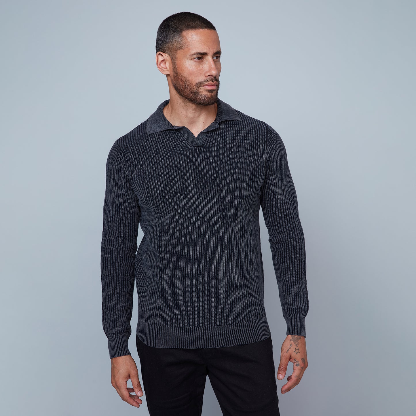 A man with short hair and a beard is standing against a plain light gray background. He is wearing a long-sleeve, textured, dark gray Johnny Collar Sweater from HEDGE, made from 100% cotton, paired with black pants. His left arm hangs by his side, revealing a tattoo on his hand and wrist, showcasing subtle fashion expertise.