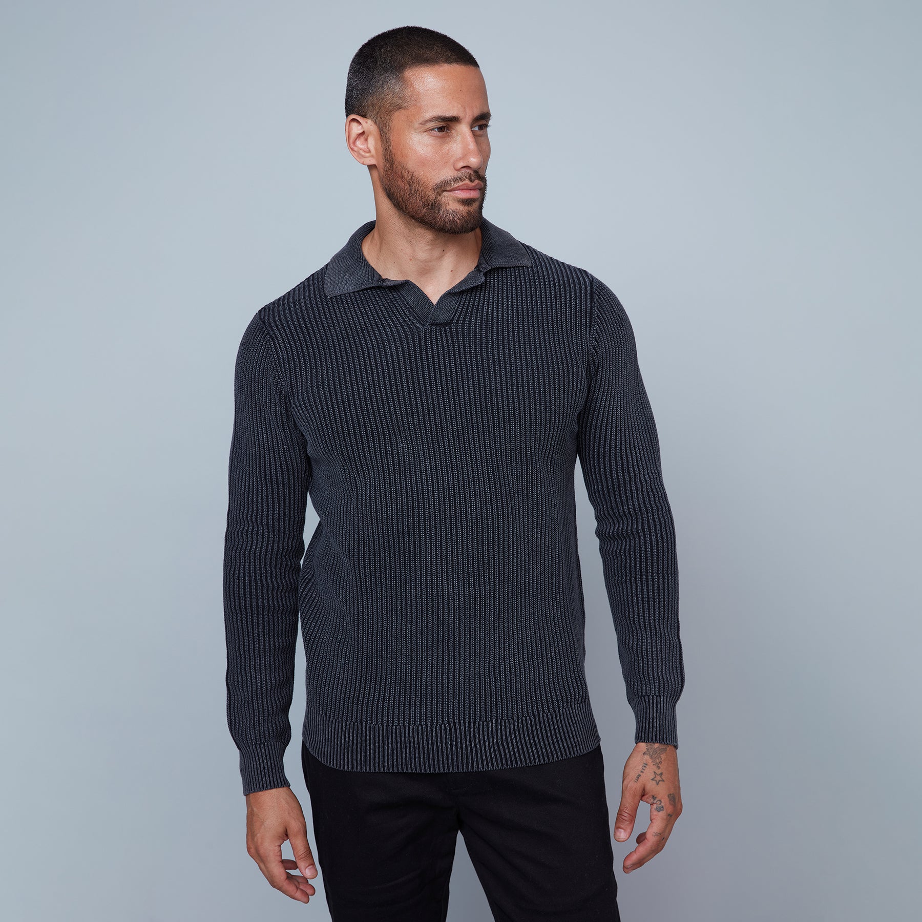 A man with short hair and a beard is standing against a plain light gray background. He is wearing a long-sleeve, textured, dark gray Johnny Collar Sweater from HEDGE, made from 100% cotton, paired with black pants. His left arm hangs by his side, revealing a tattoo on his hand and wrist, showcasing subtle fashion expertise.