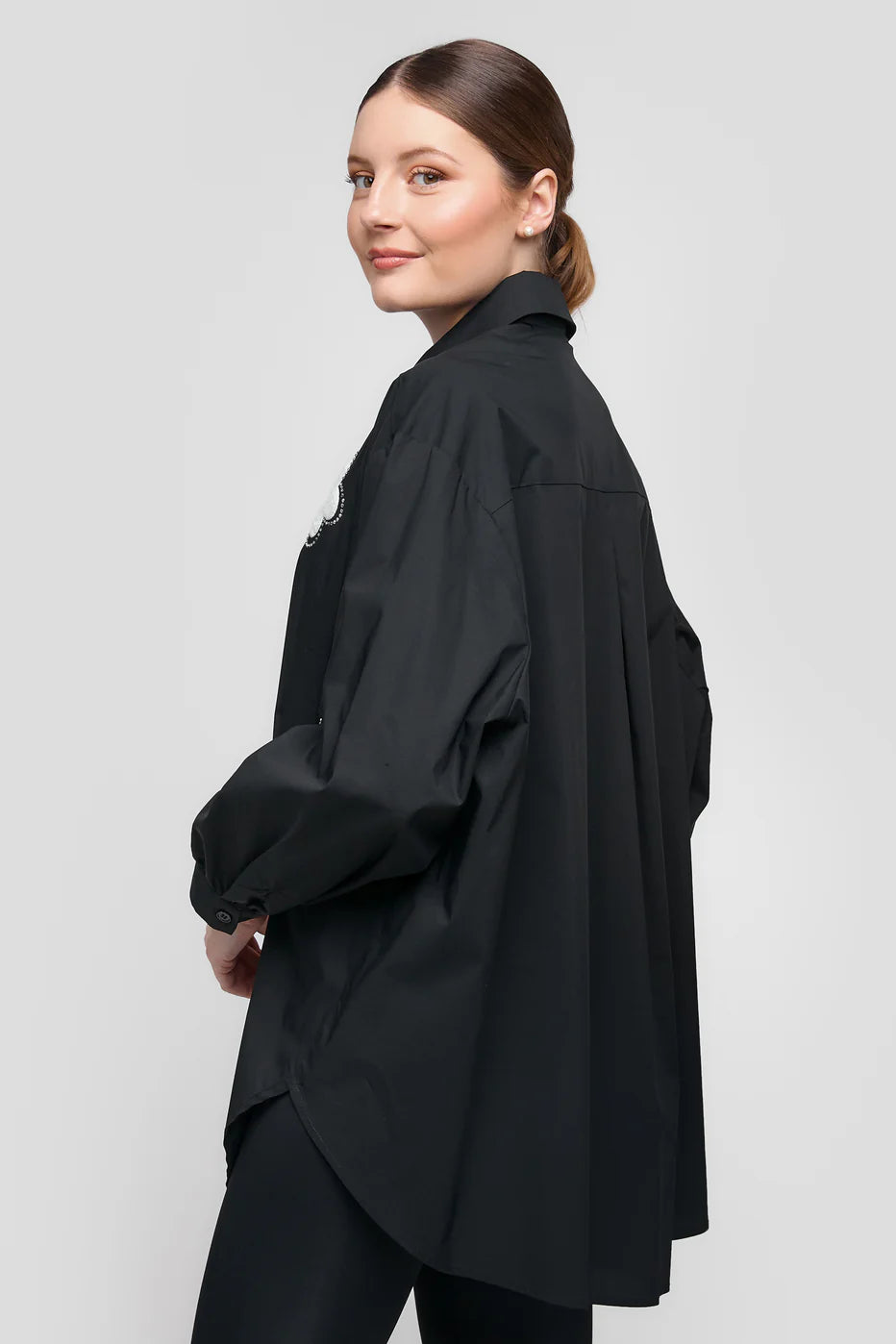 A light-skinned person in profile wears a sleek LONG SLEEVE SHIRT WITH FLORAL PATTERN CS25-600 UCHUU, paired with leggings and a bun hairstyle against a gray background. The beaded flower design on the collar is highlighted as they smile softly, showcasing elegant UCHUU Montreal style.