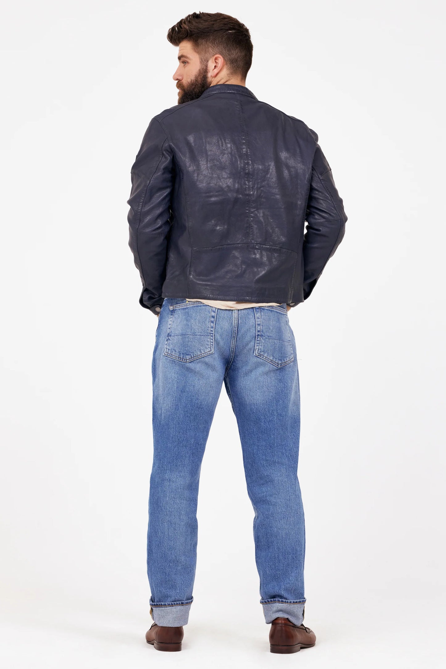 A bearded man wearing a MAURITIUS LEATHER Solvic CF Leather Jacket in navy over a white shirt, paired with light blue rolled-up jeans and brown loafers. He stands against a plain white background, holding the jacket's lapels with both hands while looking confidently towards the camera in his best-selling ensemble.