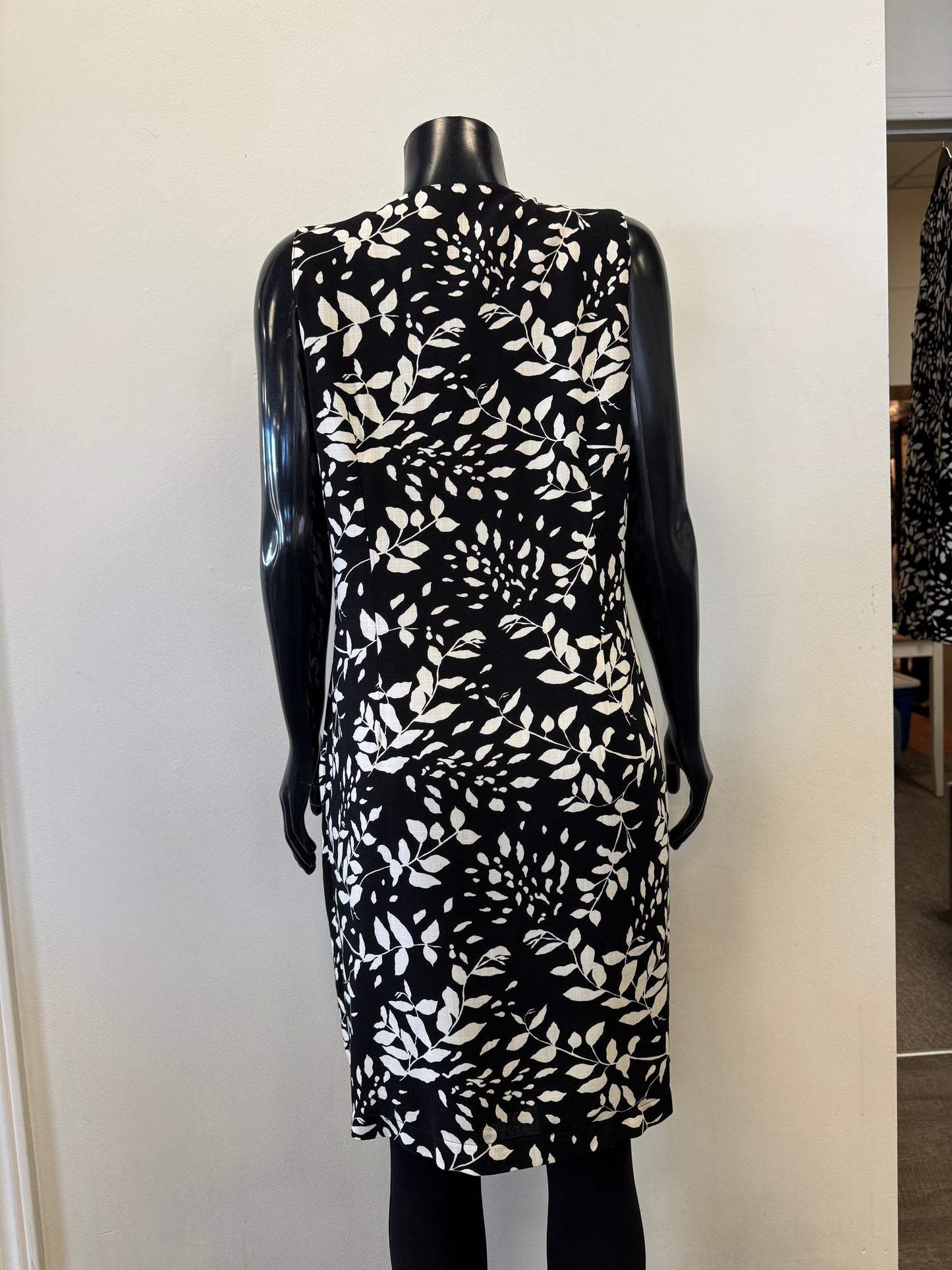 A sleek black mannequin displays Bali Corp.'s Dress 8555, a sleeveless, knee-length design featuring a white leaf pattern and crafted from 100% viscose, set against a minimal indoor backdrop.