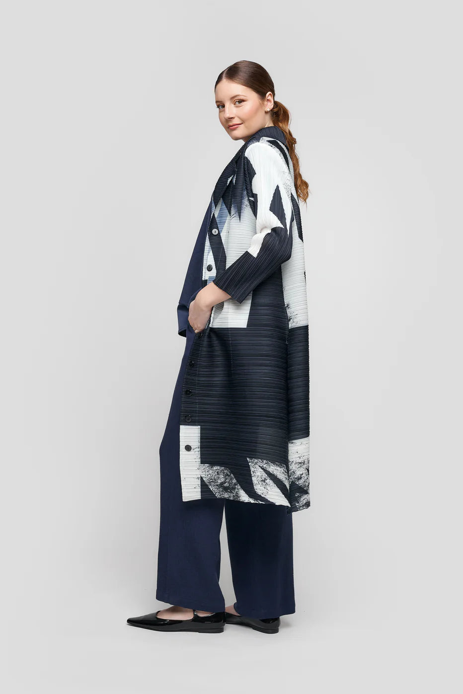 A woman models the JACKET CS25-400 UCHUU by UCHUU, a striking black and white long coat featuring abstract patterns. With her hair in a low ponytail, she pairs it with a sleek black outfit against a simple light gray background.