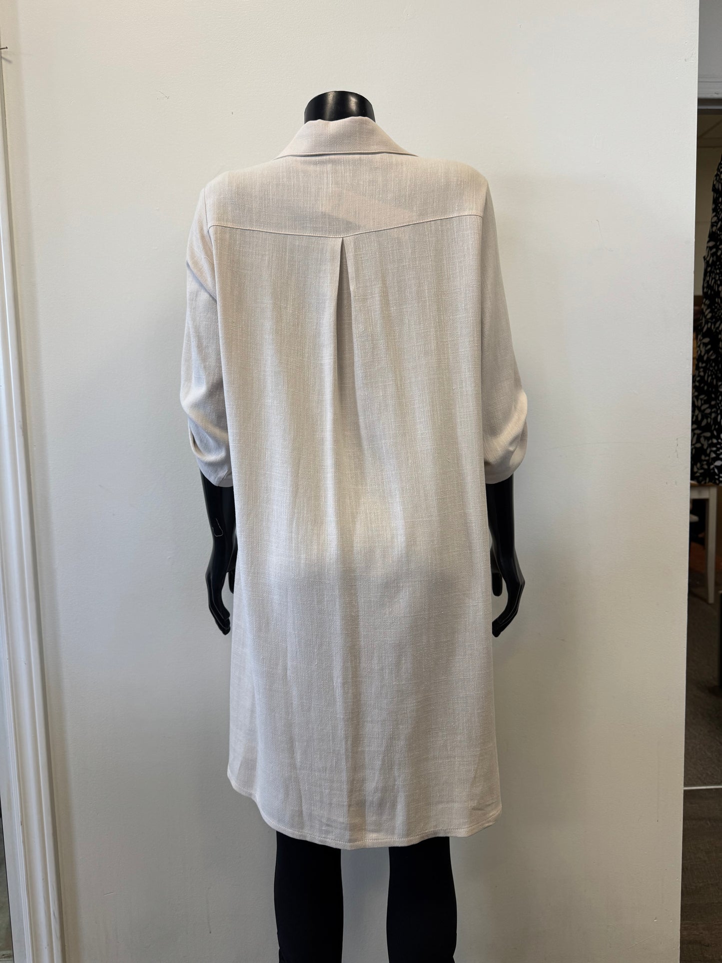 A mannequin displays Dress 8652 by Bali Corp: a long, light beige, collared shirt with rolled-up sleeves made from a Viscose and Linen blend. It features upper back pleats and falls above the knees on a plain white background with easy care instructions.