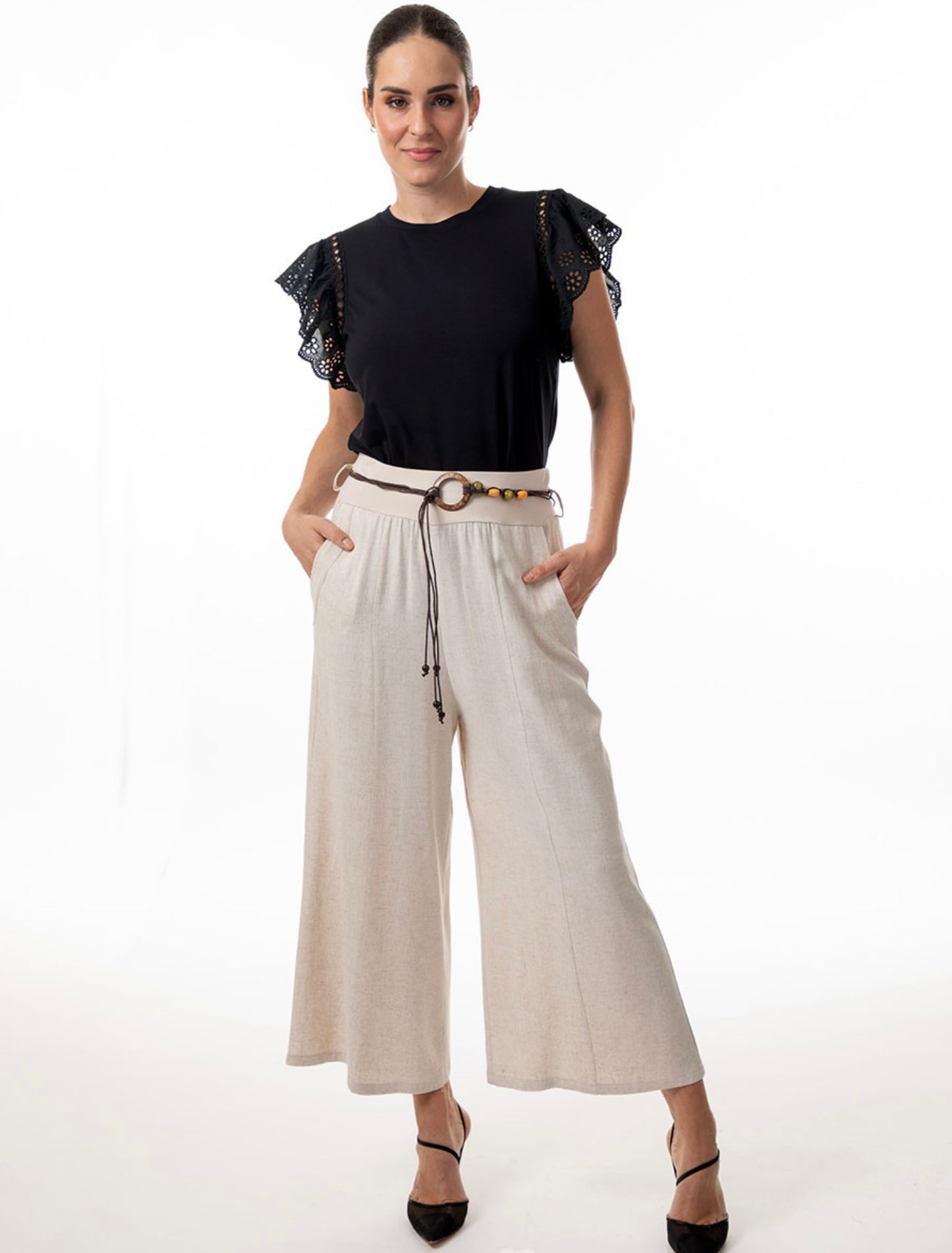 A dark-haired person is wearing a black top with decorative sleeves and light-colored, wide-leg Pant 8596 from Bali Corp, made of polyester and spandex. They stand against a white background with hands in pockets, complemented by black shoes and a belt with a decorative buckle.