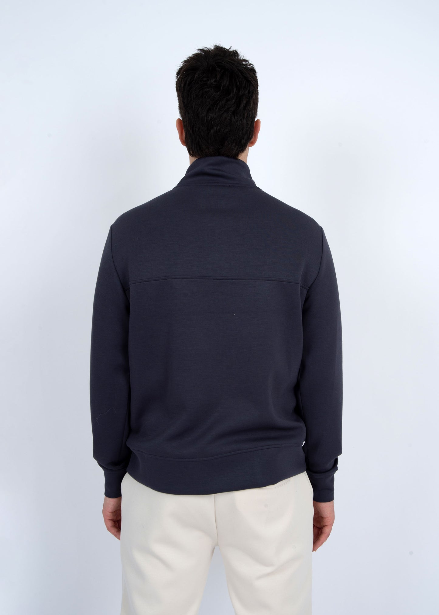A person facing away wears the Soft Knit Pullover 76MT486S2 by HEDGE, featuring a high collar and long sleeves in 100% cotton. The dark zipper sweater pairs with light pants, set against a plain white wall for stylish comfort.