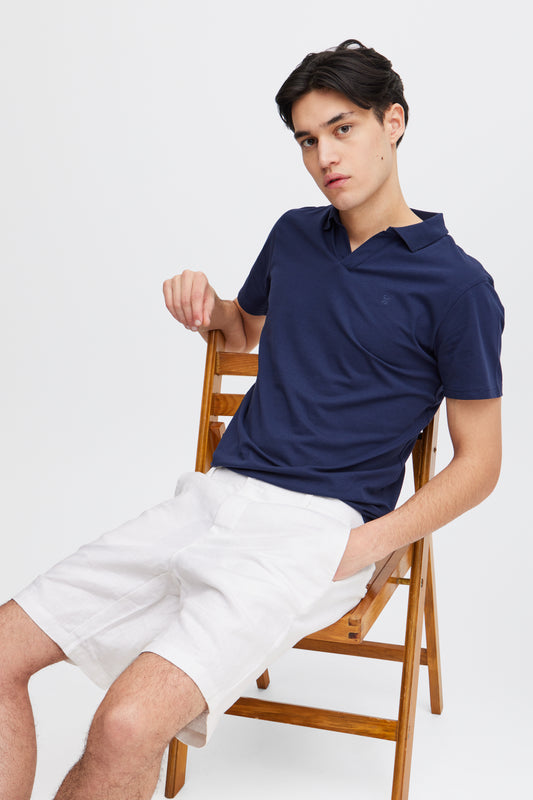 A young man with short dark hair sits on a wooden chair wearing white Linen Shorts 20505166 by Casual Fridays and a navy polo, hand in pocket, looking relaxed—an effortless style perfect for casual Friday comfort.