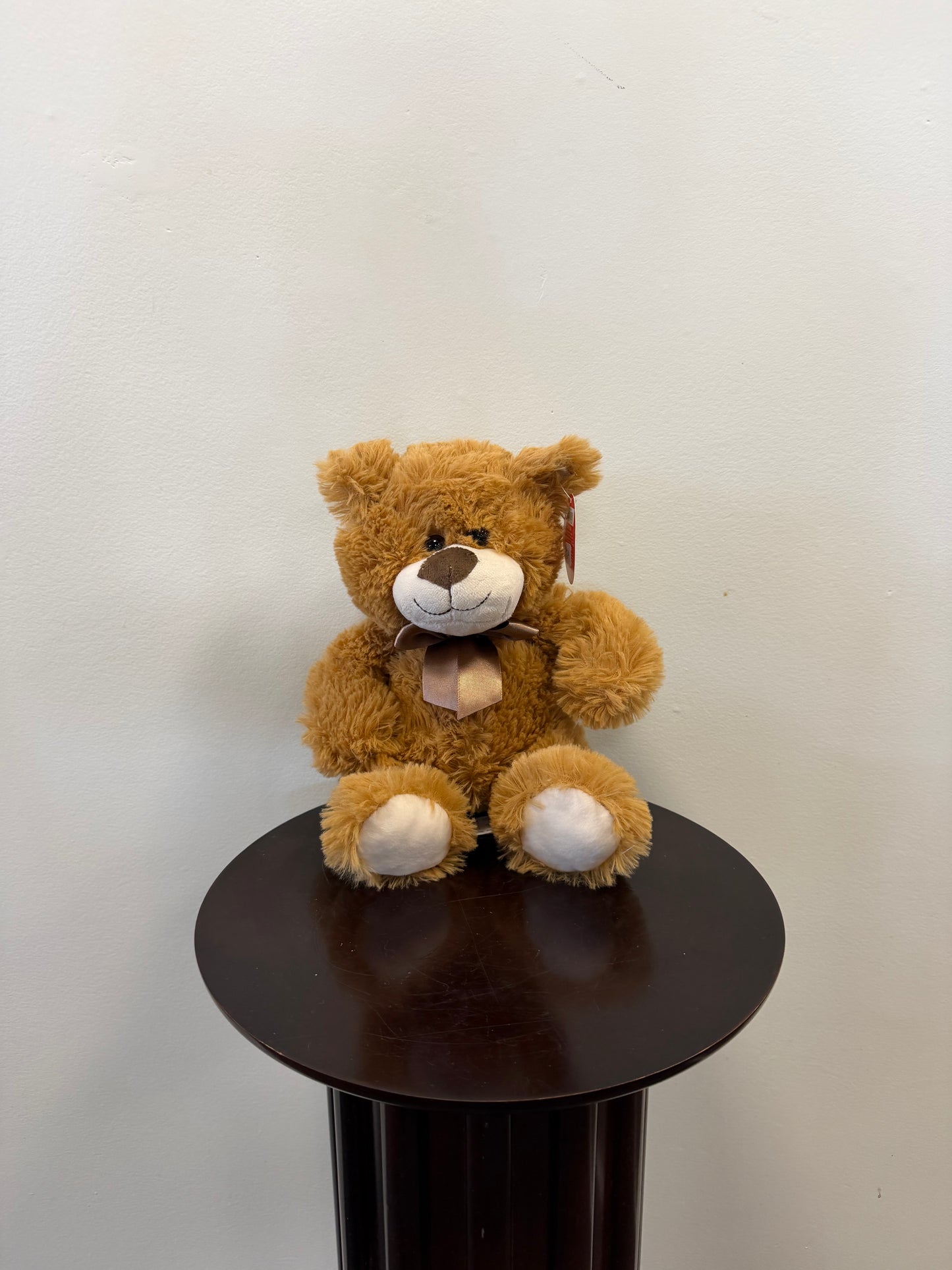 A Classic Teddy Bear – Fiesta by Fiesta, featuring a beige snout and a bow, sits on a dark round pedestal against a plain light-colored wall—making it a charming choice for gift delivery.