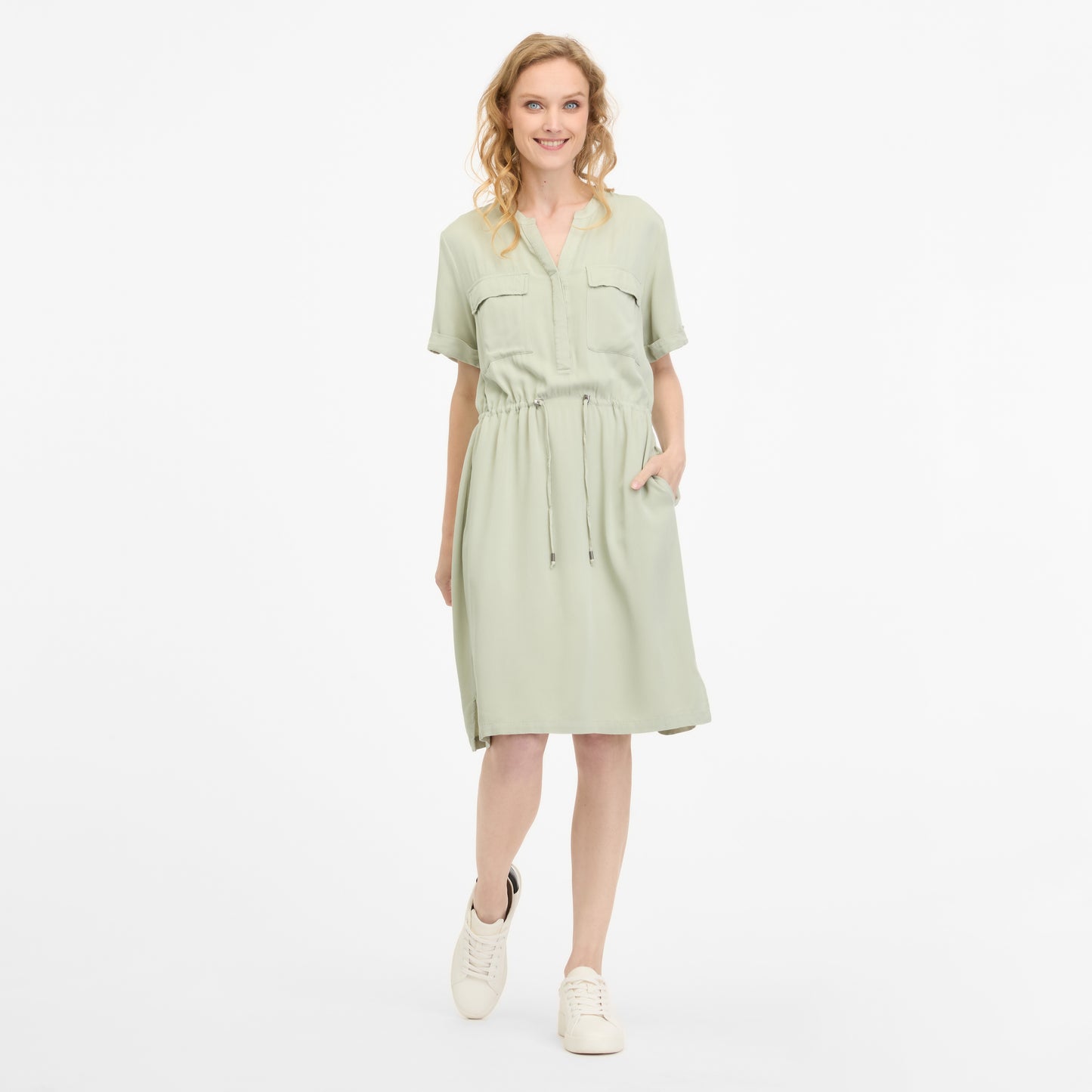 A woman in a light green ROISANA DRESS 2511-20026-5036 by RAGWEAR smiles with one hand in her pocket. The dress, made from 100% viscose, has a drawstring waist and short sleeves. She pairs it with white sneakers, and her wavy blonde hair stands out against the plain white background.