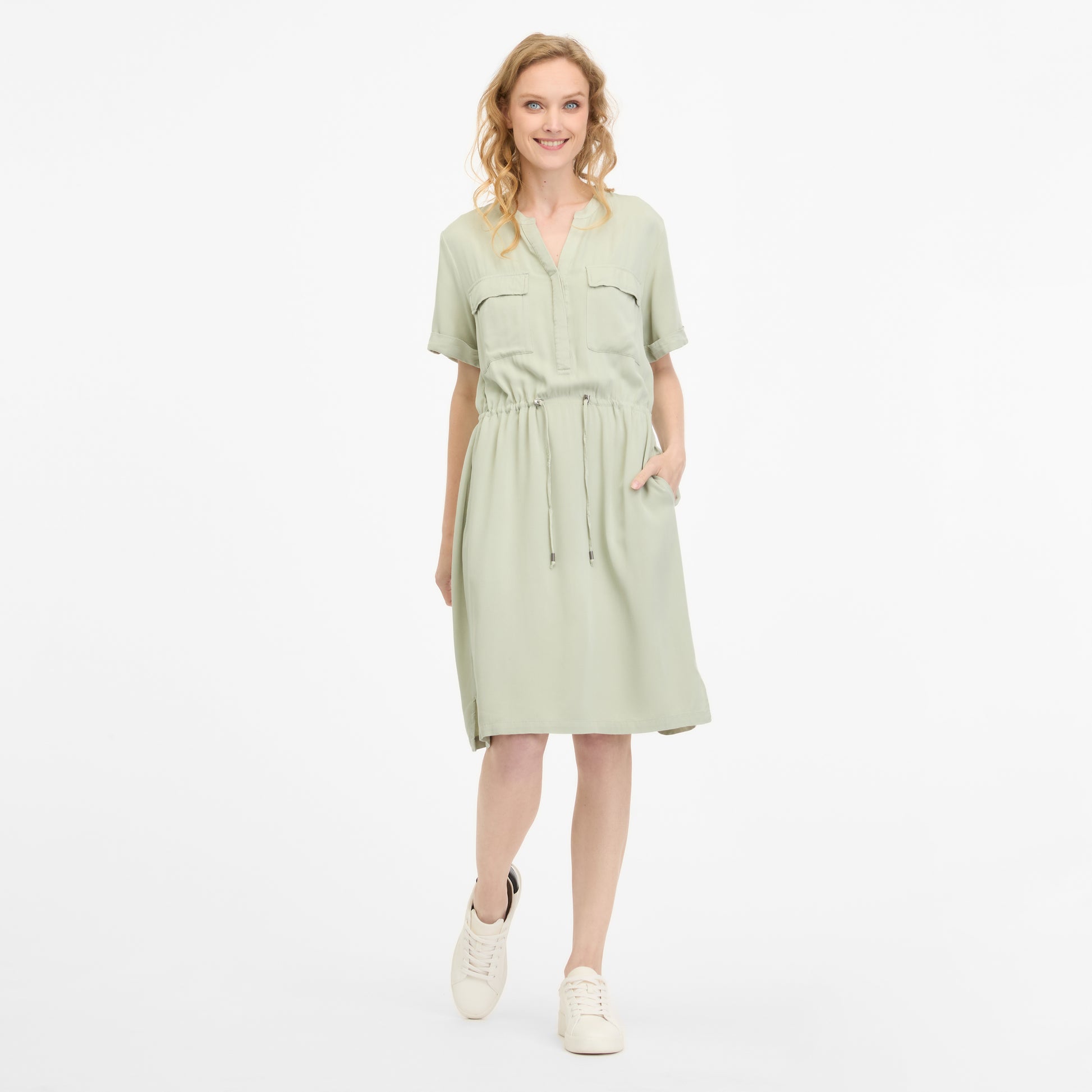 A woman in a light green ROISANA DRESS 2511-20026-5036 by RAGWEAR smiles with one hand in her pocket. The dress, made from 100% viscose, has a drawstring waist and short sleeves. She pairs it with white sneakers, and her wavy blonde hair stands out against the plain white background.