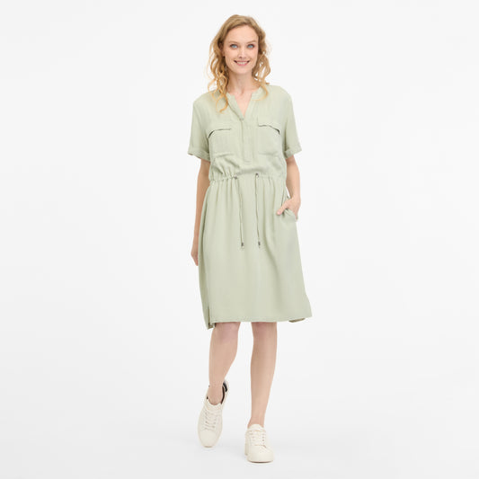 A woman in a light green ROISANA DRESS 2511-20026-5036 by RAGWEAR smiles with one hand in her pocket. The dress, made from 100% viscose, has a drawstring waist and short sleeves. She pairs it with white sneakers, and her wavy blonde hair stands out against the plain white background.