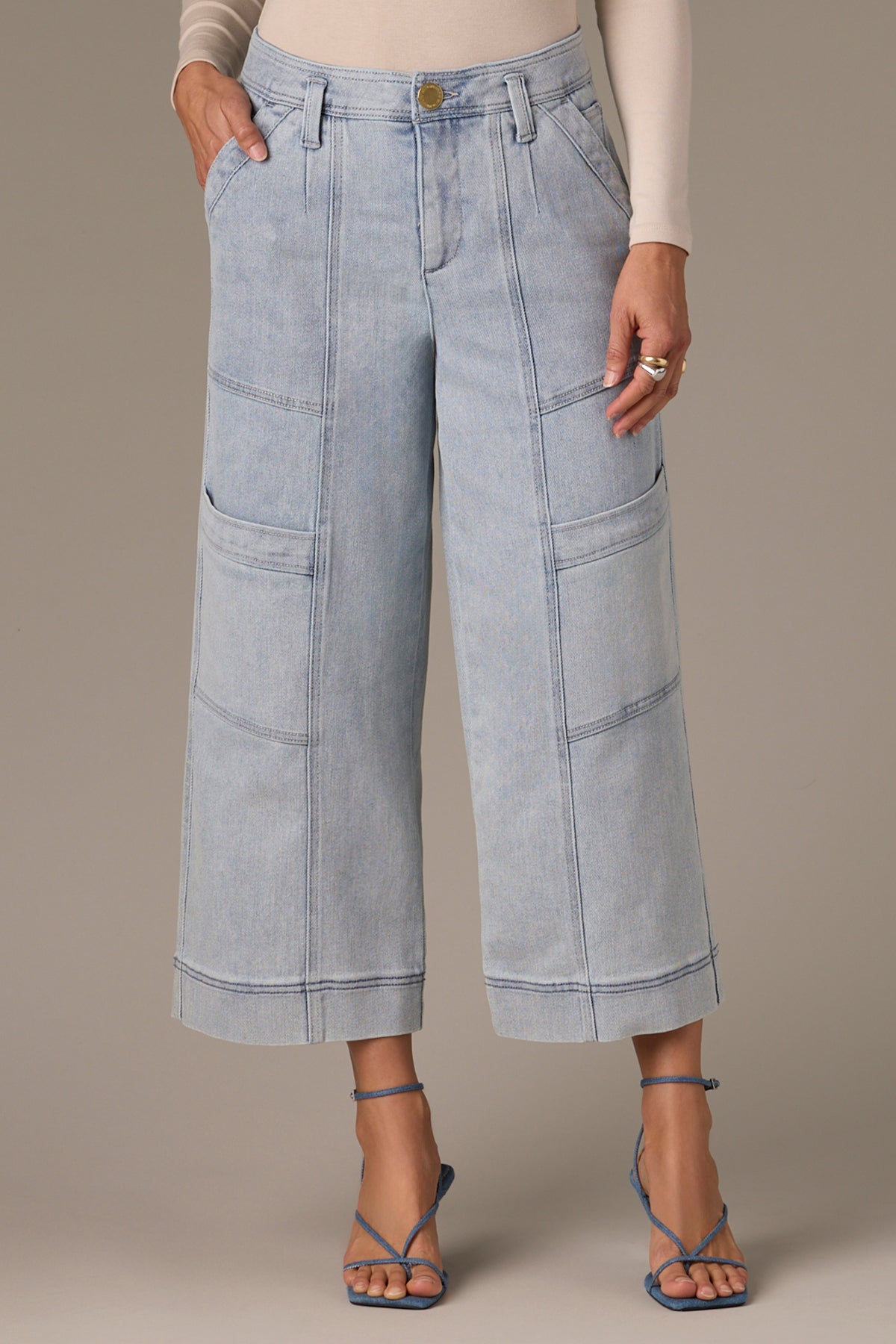 Wearing Skyrise Double Bottom Wide Leg Jeans by Wit & Wisdom, a person stands with hands in pockets. The light denim, high-waisted jeans feature large pockets and are paired with a beige long-sleeve top and blue strappy sandals against a plain grayish background.