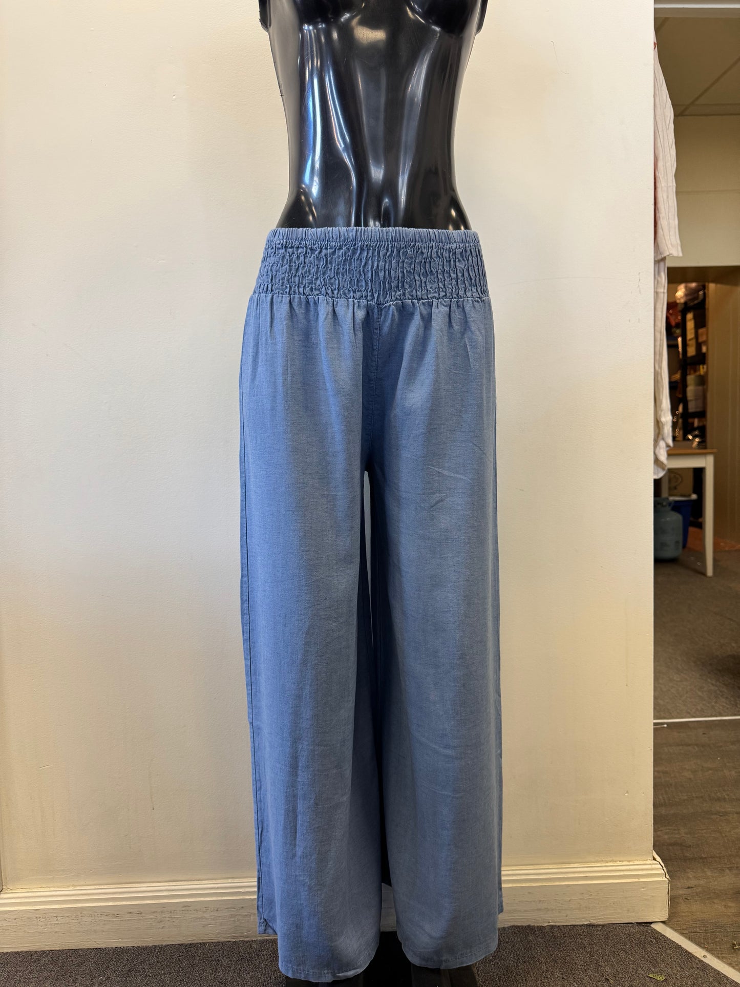 A black mannequin displays Bella Amore's Linen Blend Pants 623, featuring light blue wide legs and an elastic waistband, epitomizing Italian style and comfort. The plain off-white backdrop offers a glimpse of the store's interior on the right.