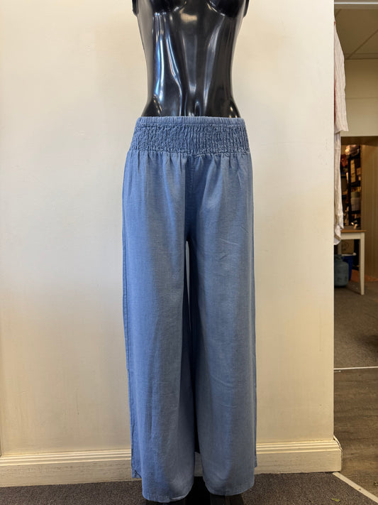 A black mannequin displays Bella Amore's Linen Blend Pants 623, featuring light blue wide legs and an elastic waistband, epitomizing Italian style and comfort. The plain off-white backdrop offers a glimpse of the store's interior on the right.
