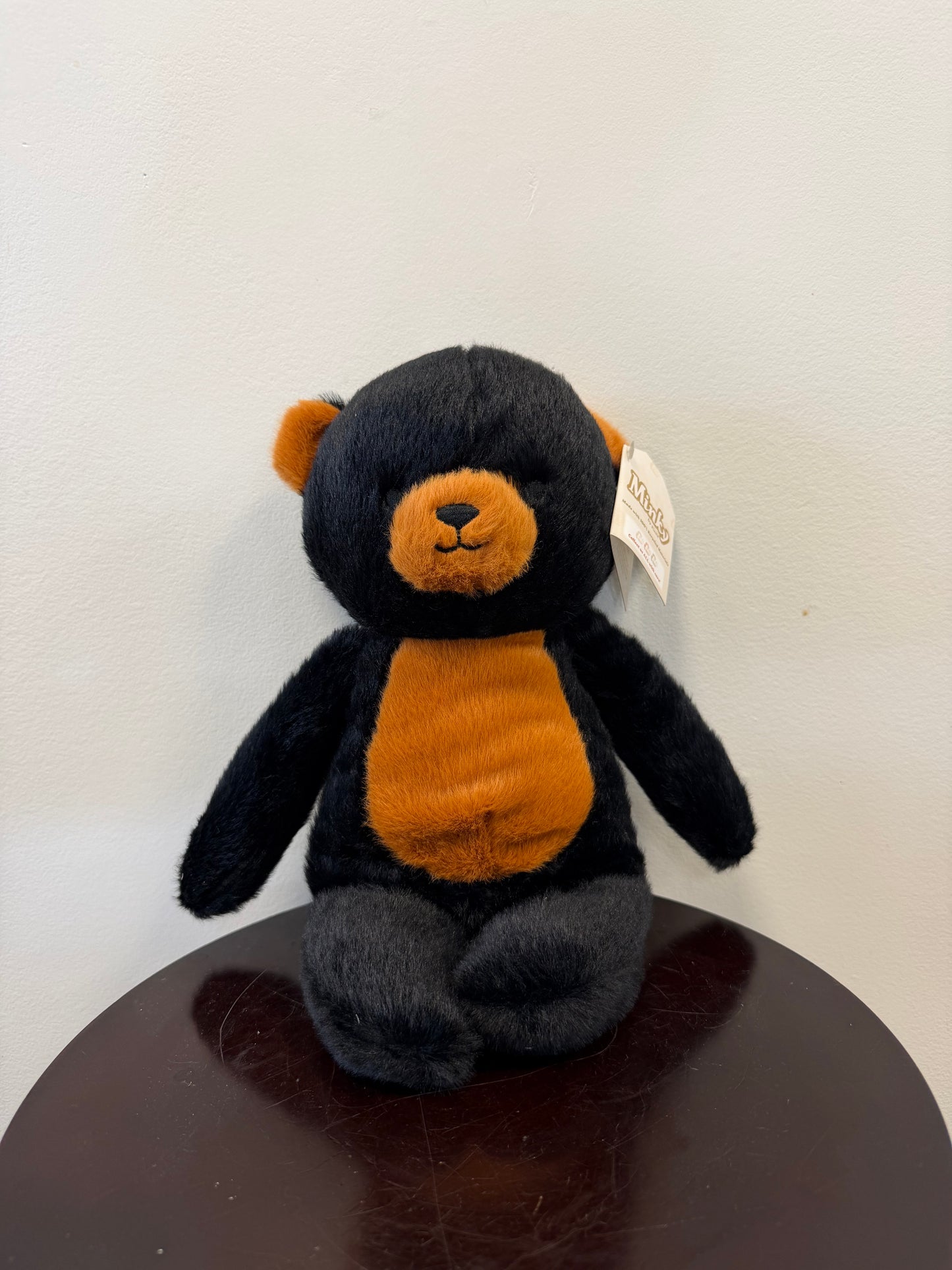 Balto the Black Bear – Minky by Fiesta, an eco-friendly stuffed animal made from recycled materials, sits upright on a dark round table against a plain white wall. This plush bear by Fiesta features soft black fur with orange accents on its snout, chest, and inner ears, and has a small tag attached to its left ear.