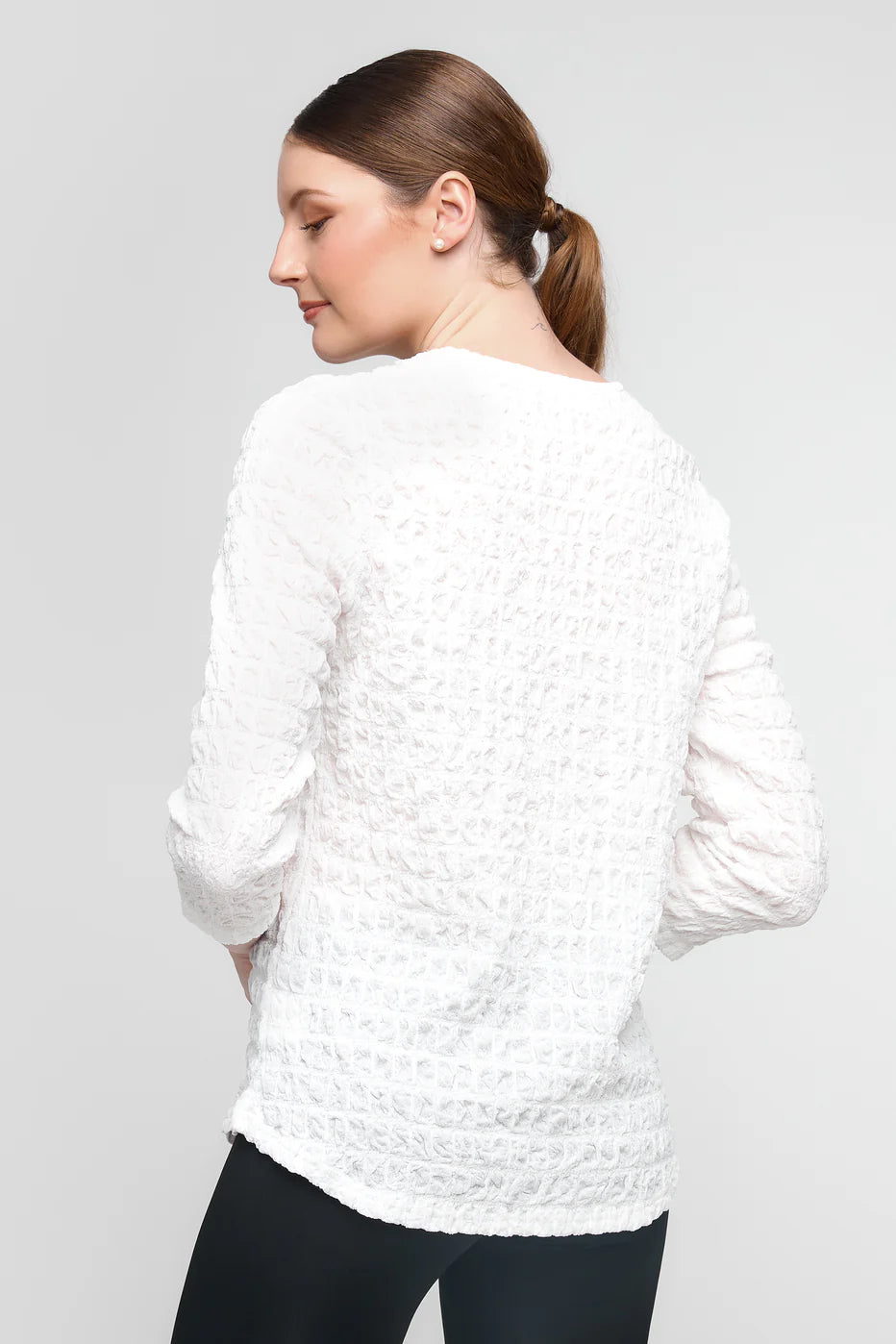 A woman with long brown hair in a ponytail wears a textured white, long-sleeved polyester sweater, paired with black pants. Facing sideways and looking down, the plain light gray background highlights the comfort of her UCHUU-inspired LONG-SLEEVED CREW NECK T-SHIRT CS25-308 ensemble.