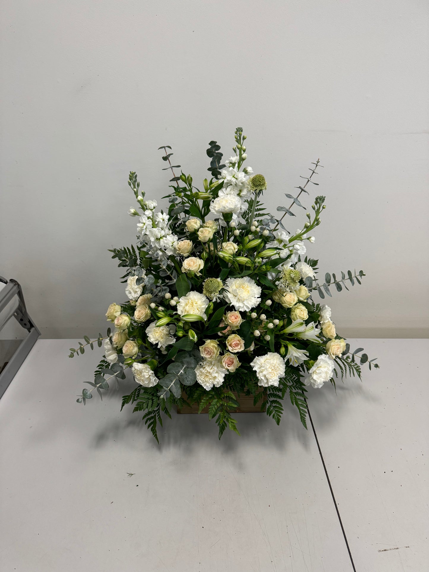The White Sympathies Arrangement from Posies Flowers & Fashion features a wooden container that holds a lush assortment of white and cream flowers, including roses, carnations, and chrysanthemums. Green foliage and eucalyptus leaves extend gracefully from the sides, set against a plain white background. Please check availability for this elegant floral display.