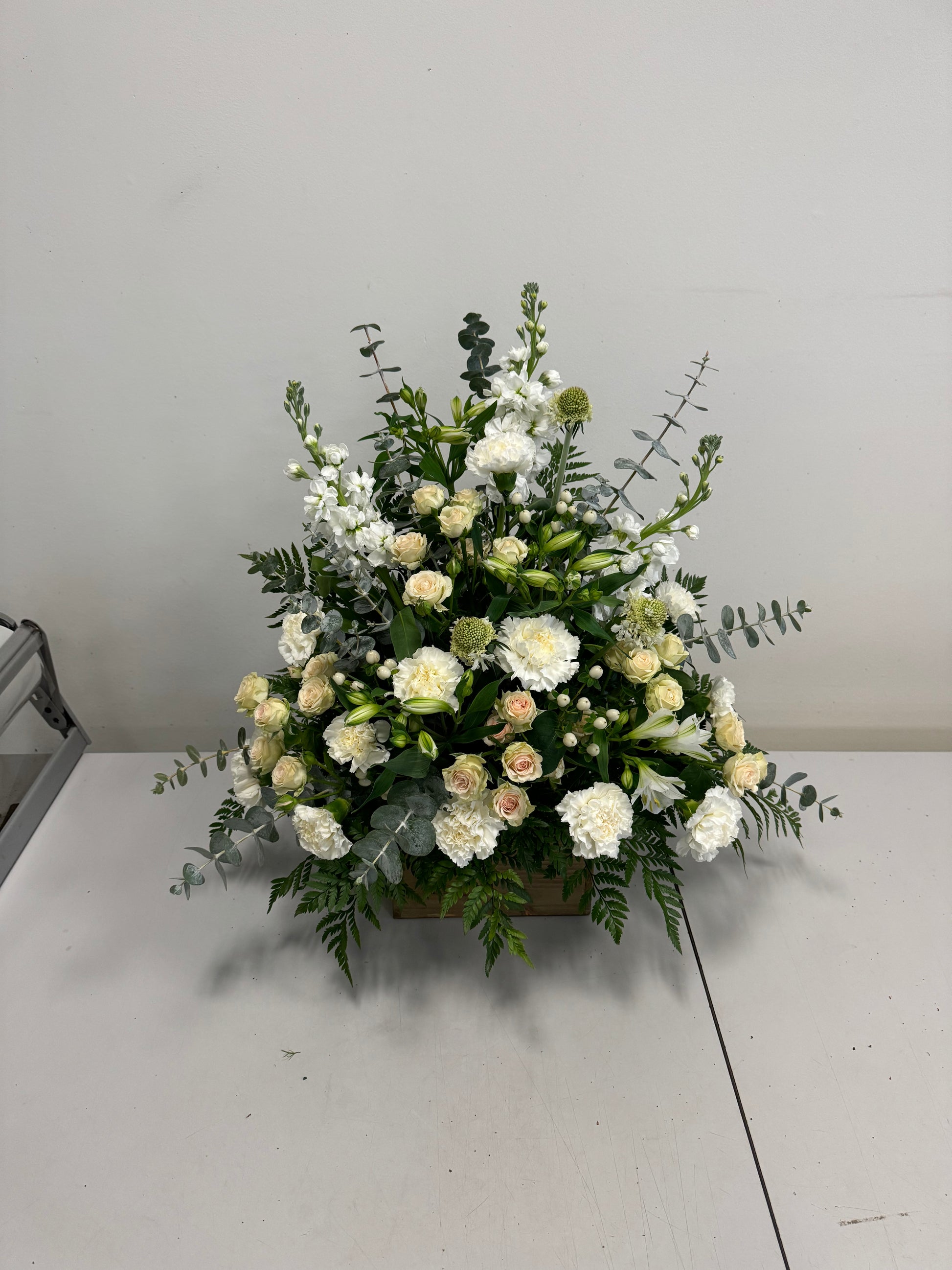 The White Sympathies Arrangement from Posies Flowers & Fashion features a wooden container that holds a lush assortment of white and cream flowers, including roses, carnations, and chrysanthemums. Green foliage and eucalyptus leaves extend gracefully from the sides, set against a plain white background. Please check availability for this elegant floral display.